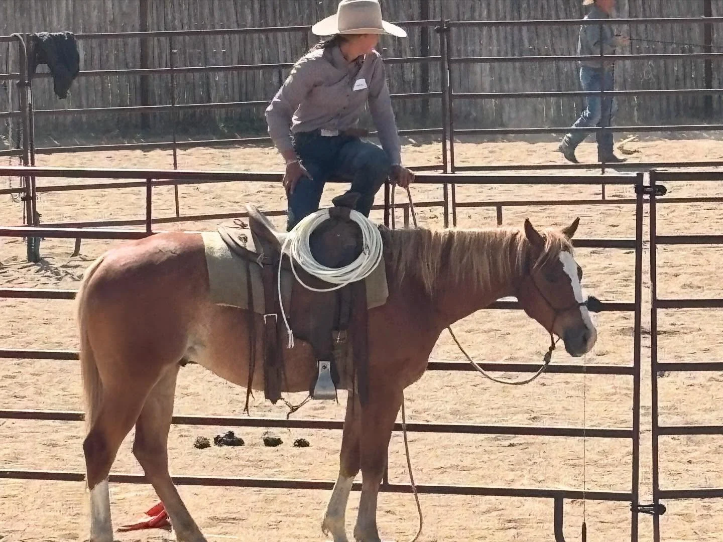 A person wearing a cowboy hat appears to be getting on a horse in an outdoor, sandy arena. The person is dressed in a long-sleeve shirt and jeans, holding a coiled lasso, with a fence surrounding the area.
