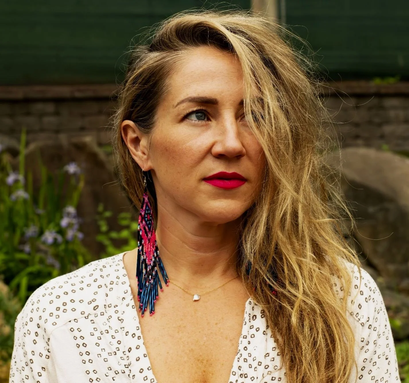 Woman with long hair and red lipstick wearing a multi-colored earring and a white patterned blouse.