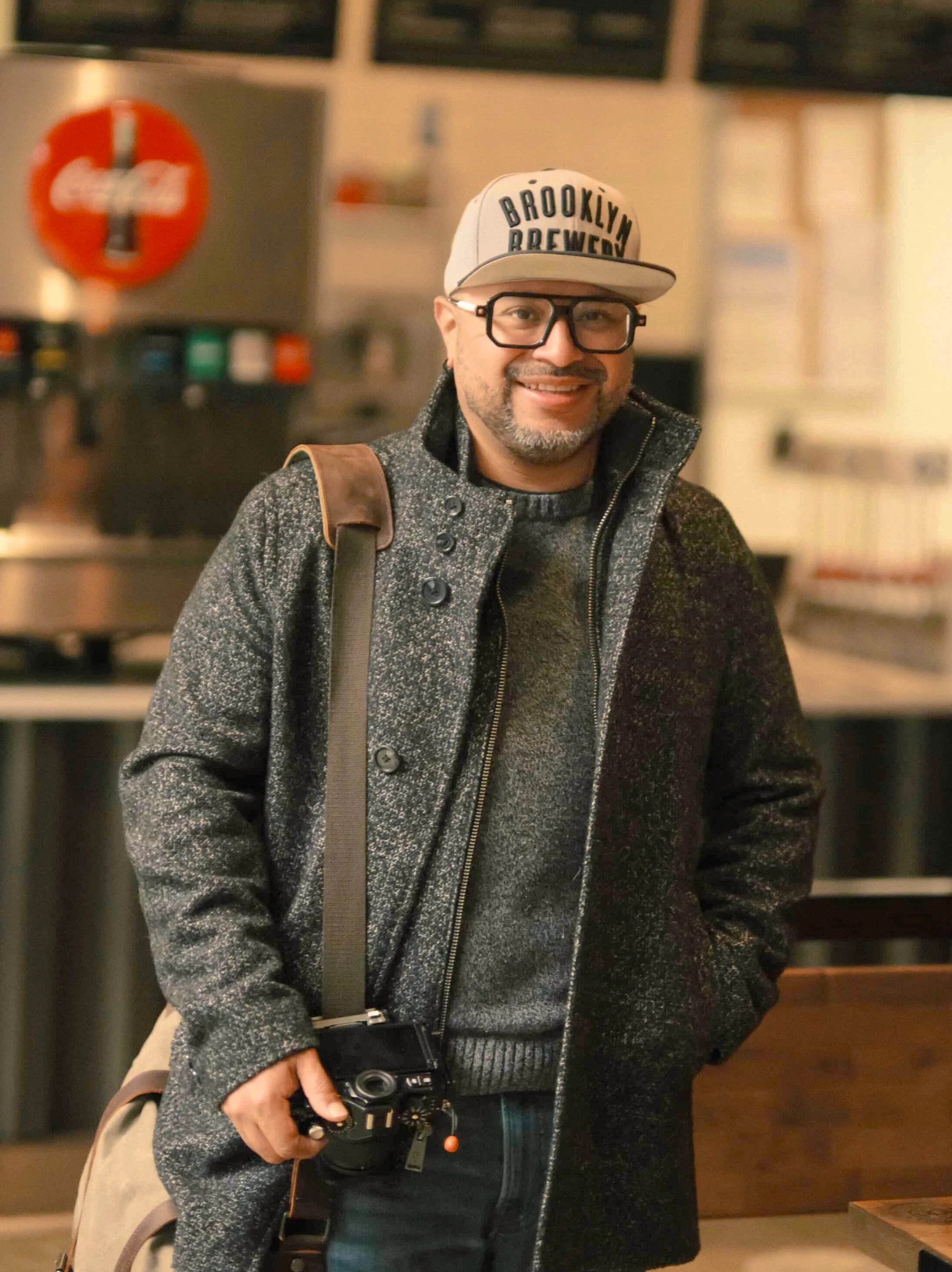 Portrait of a photographer wearing a grey wool coat, Brooklyn Brewery baseball cap, and thick-framed glasses holding a camera in a café setting.
