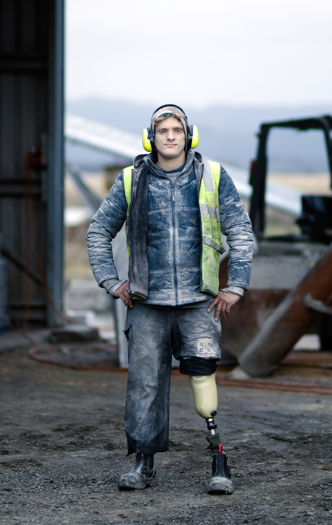 Matt in his farm outfit with construction vest and headphones with pant leg rolled up showing his prostethic leg with forklift in background