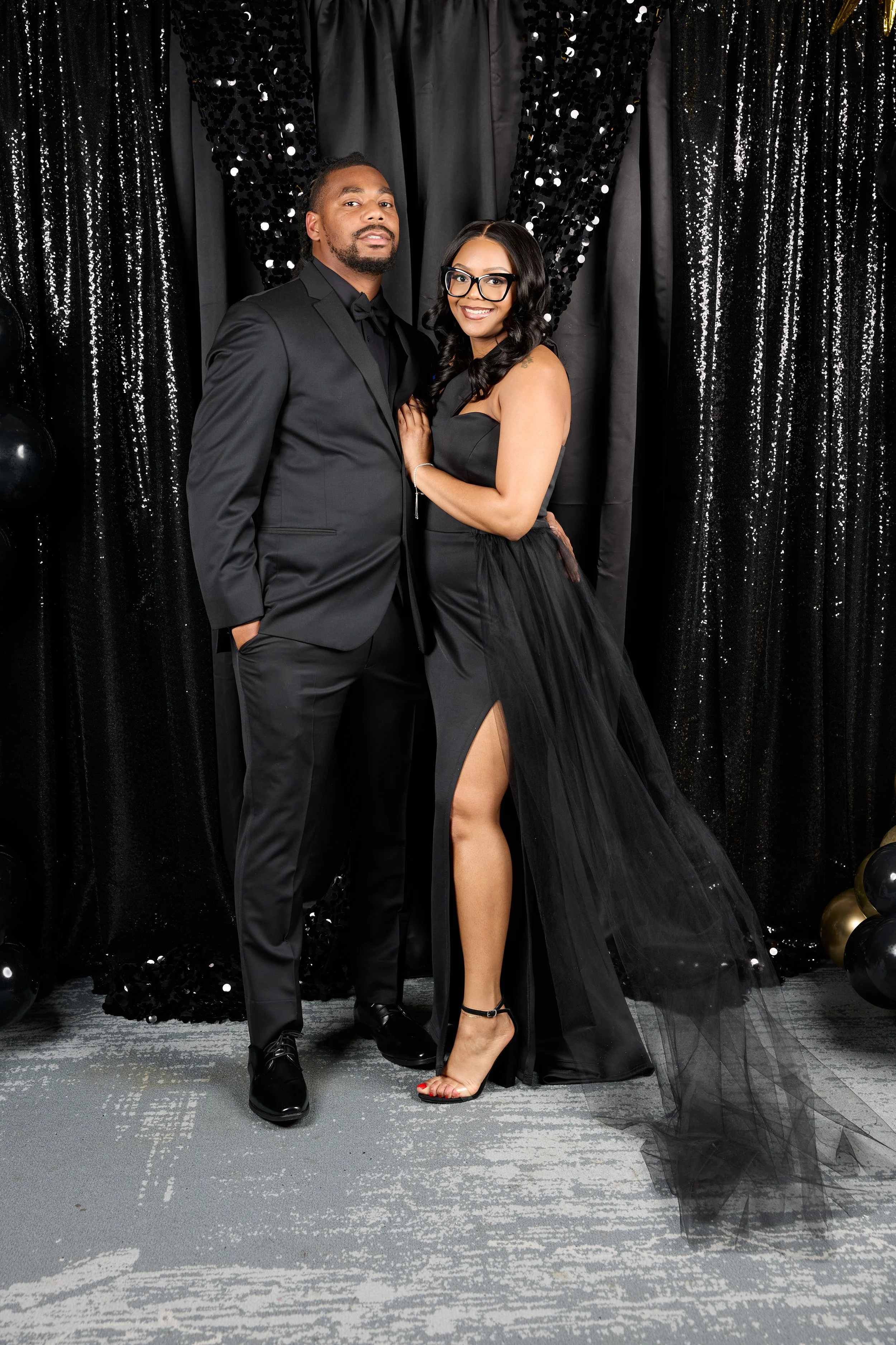 A man and woman dressed in formal black attire standing together against a black backdrop with sequins and balloons.