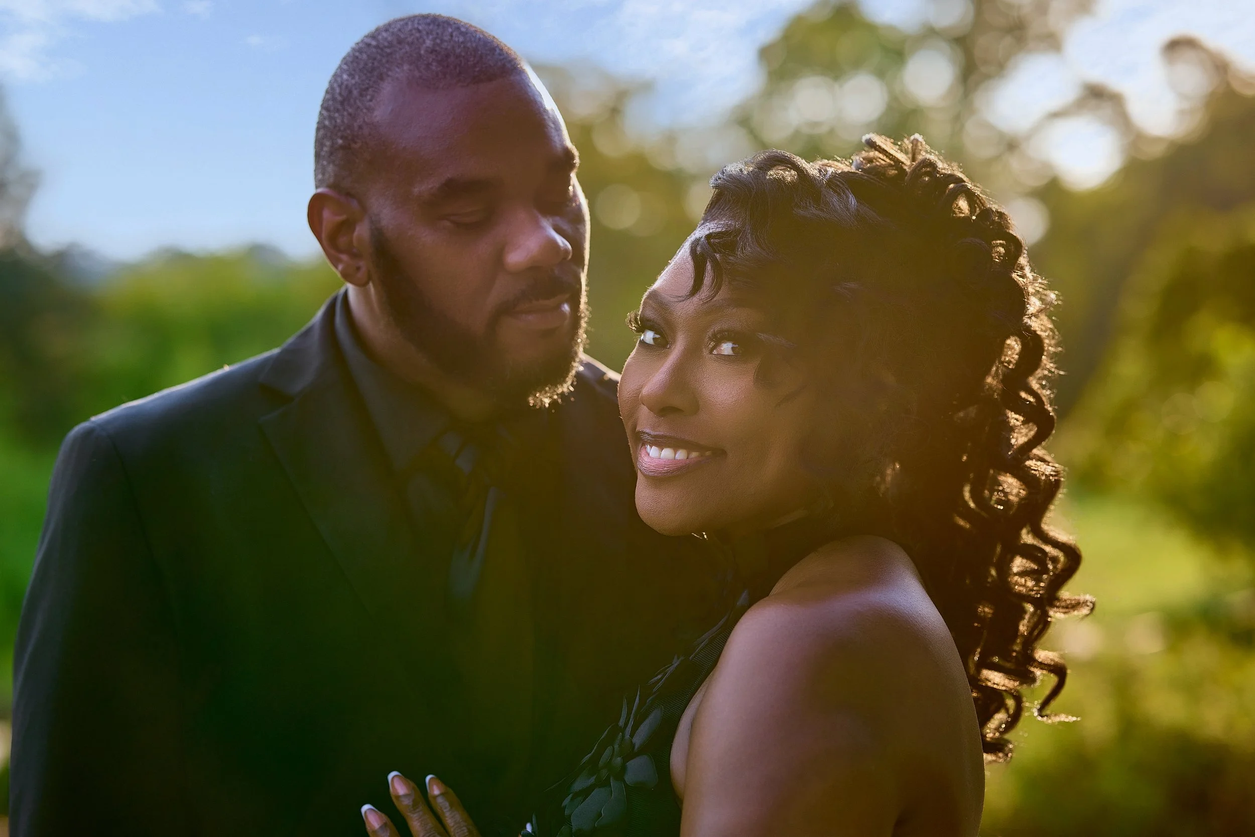 A man and woman standing close together outdoors on a sunny day, with the woman smiling and the man looking down at her.