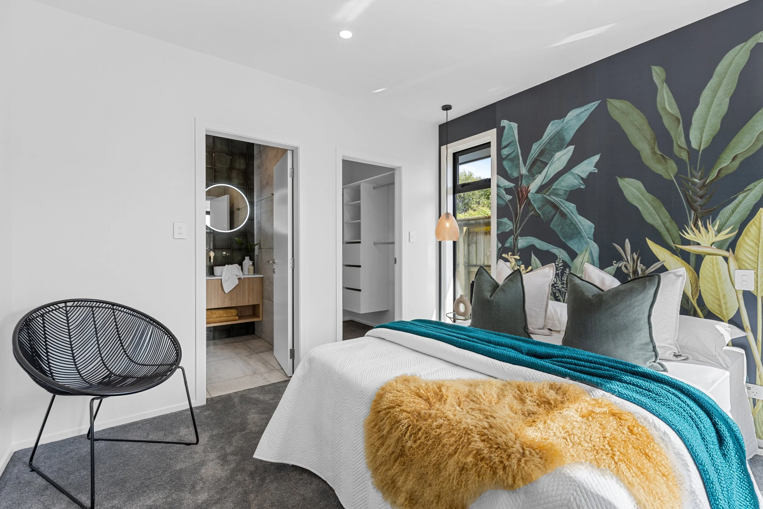 Modern bedroom with tropical wallpaper accent wall, white bedding, decorative pillows, teal throw, yellow faux fur blanket, black wire chair, window with view of greenery, bathroom with round mirror, and walk-in closet.