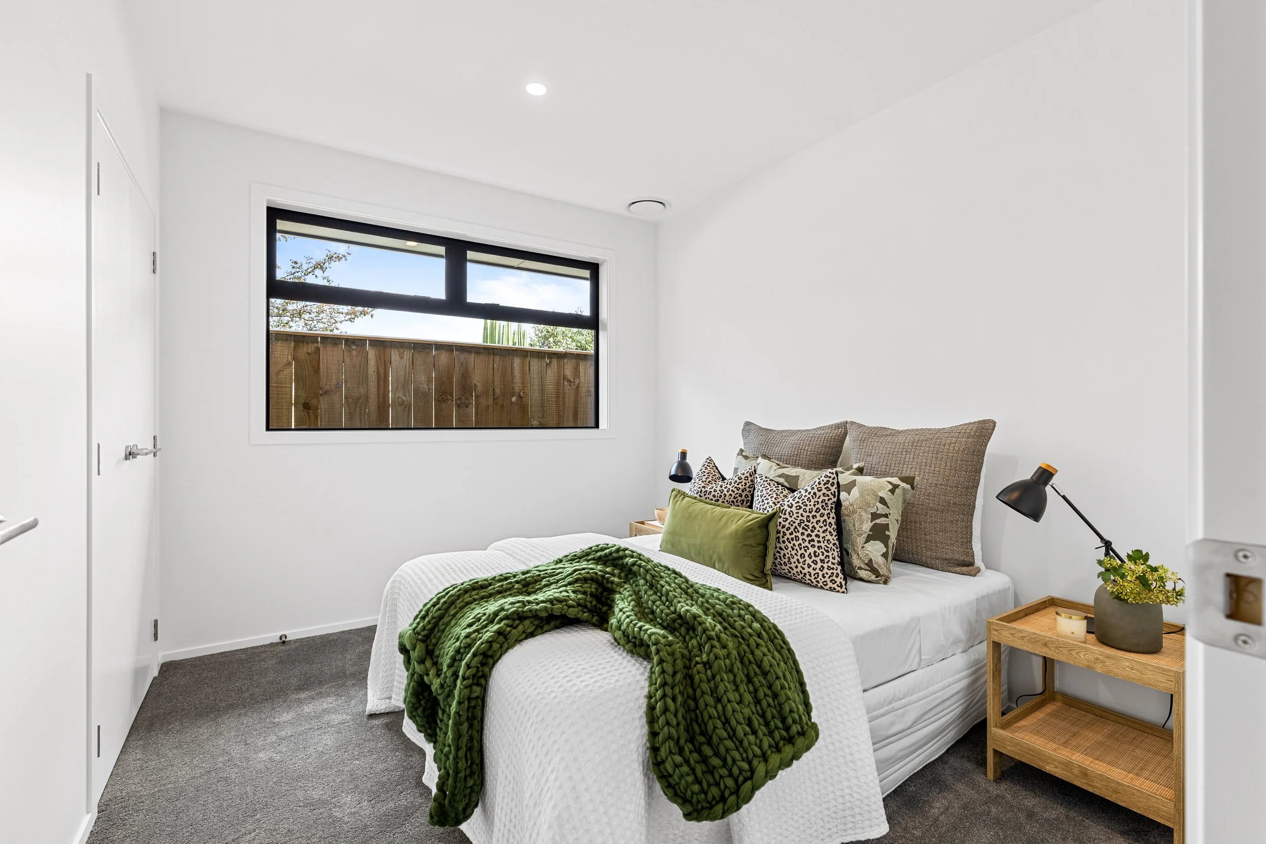 Bedroom with white walls, a large window, a bed with multiple pillows, a green chunky knit blanket, and a wooden nightstand with a black lamp, candle, and plant.