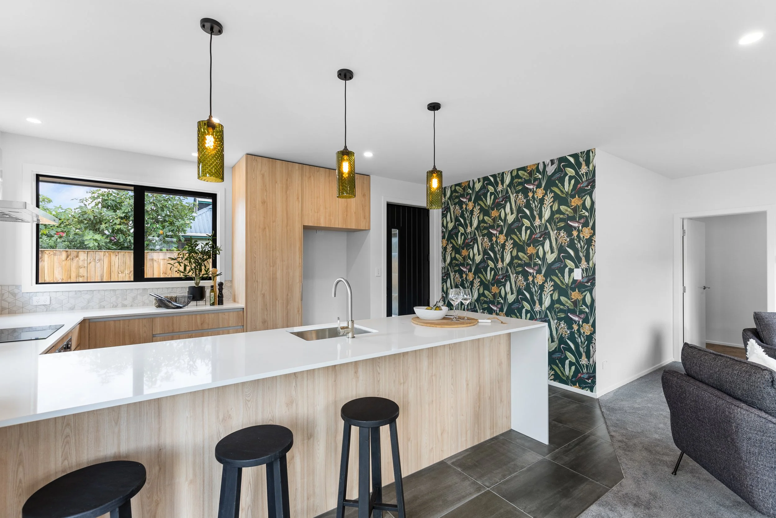 Modern kitchen with light wood cabinetry, white island counter with a sink, three black stools, and light fixtures hanging above. A window shows greenery outside, and there is a decorative wallpapered accent wall.
