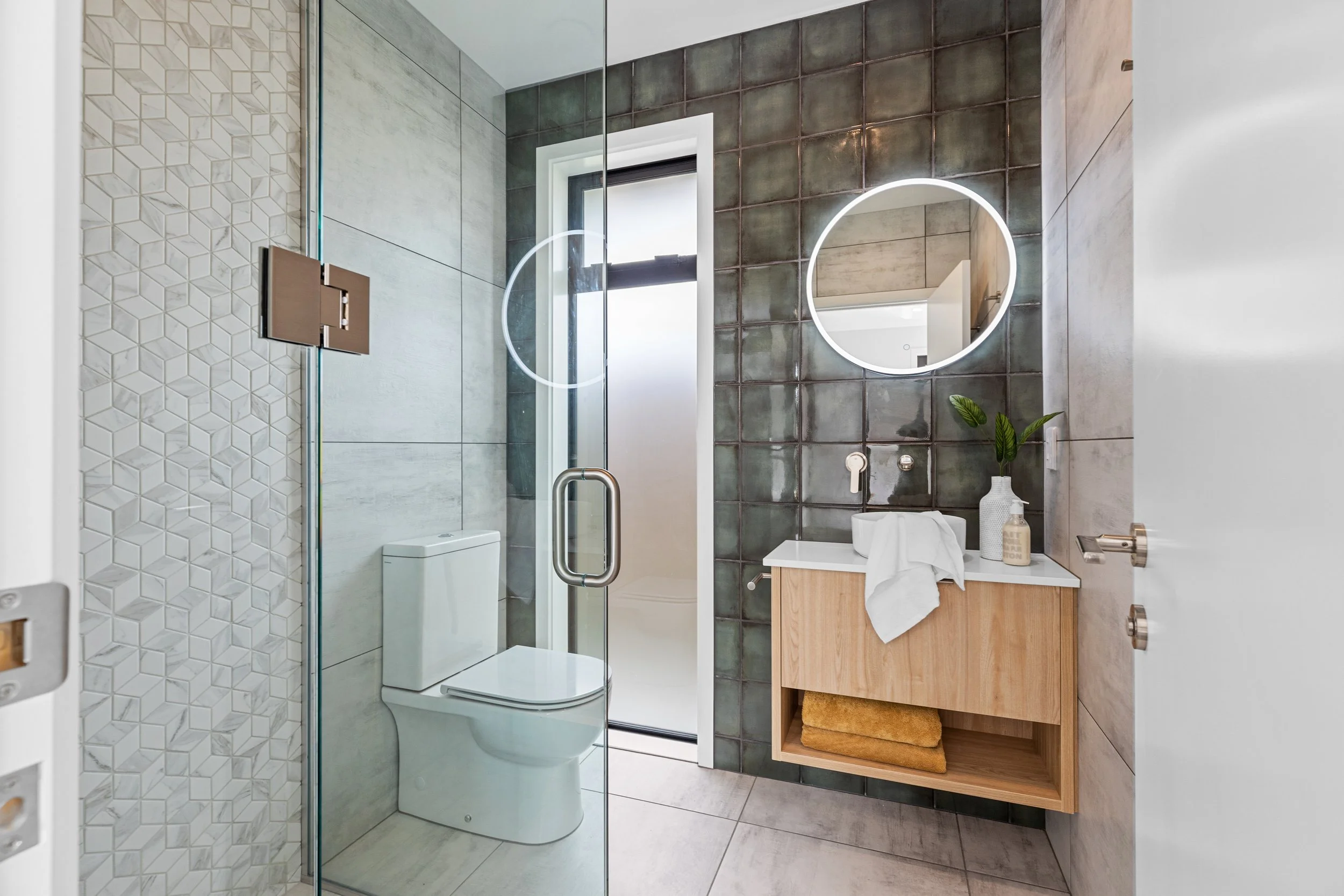 Modern bathroom interior with a glass shower enclosure, toilet, wooden vanity with a round mirror, potted plant, and neatly folded towel.