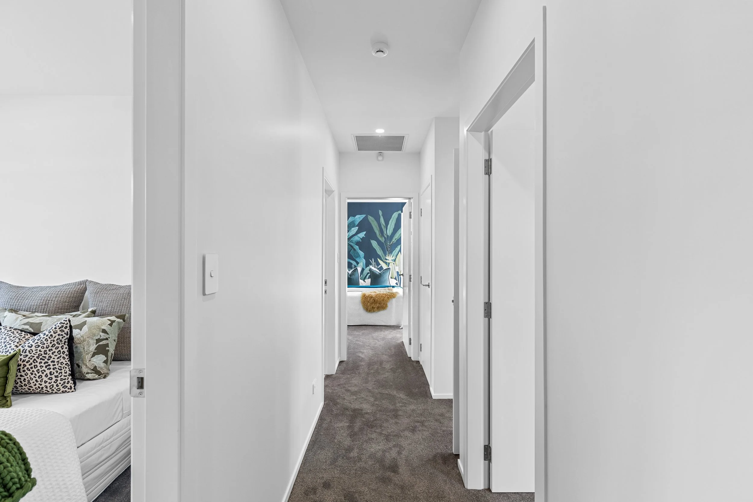 Cozy hallway with white walls leading to a bedroom with large plant wall art, cluttered with a bed, pillows, and blanket.
