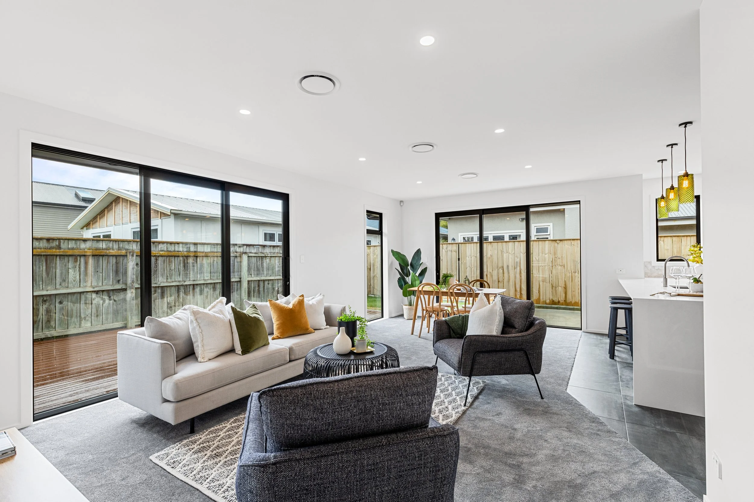 Modern living room with white walls, grey carpet, and large sliding glass doors leading to a backyard. Contains white sofa with colorful pillows, dark armchairs, a black coffee table with vases, a dining table with wooden chairs, and kitchen area wit