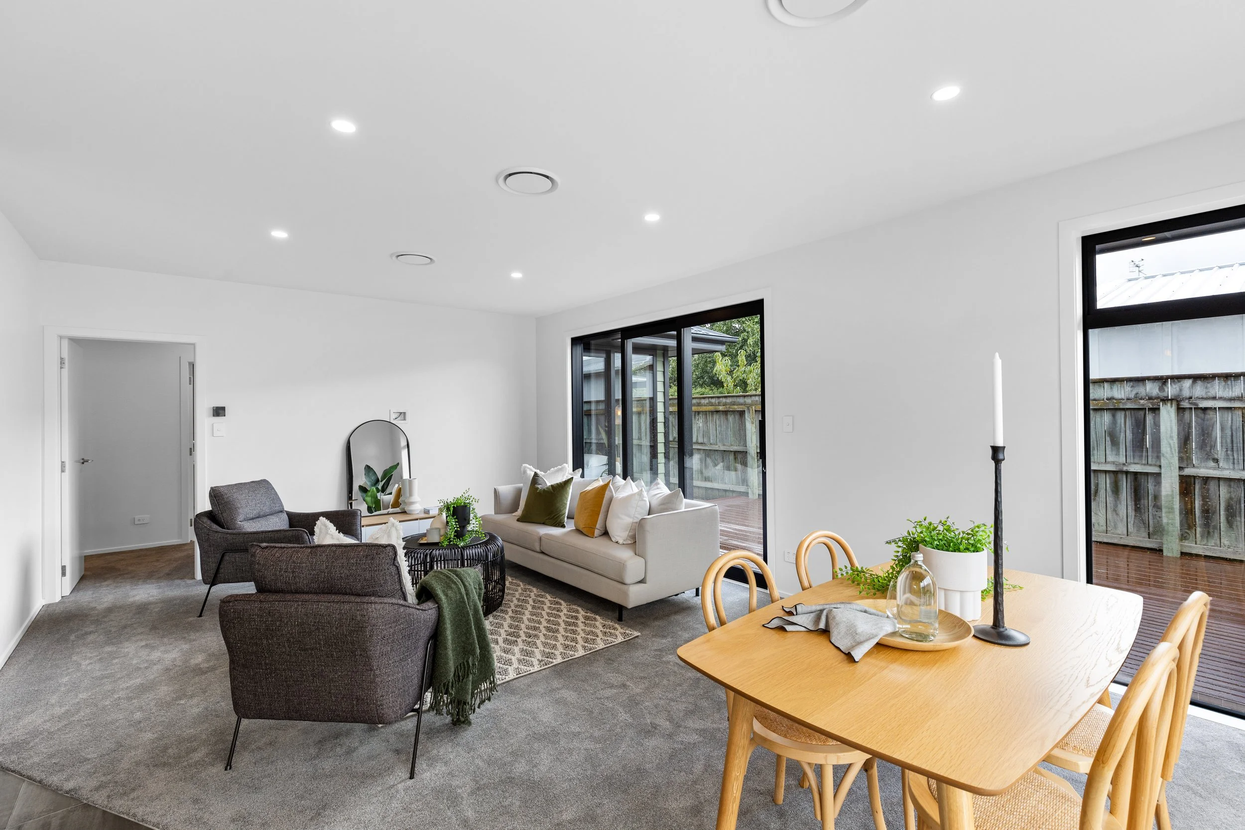 Living room with white walls, gray carpet, and modern furniture. There is a white sofa with colorful pillows, two gray armchairs, a black coffee table, and a wooden dining table with chairs. Large sliding glass doors open to an outdoor deck. The room