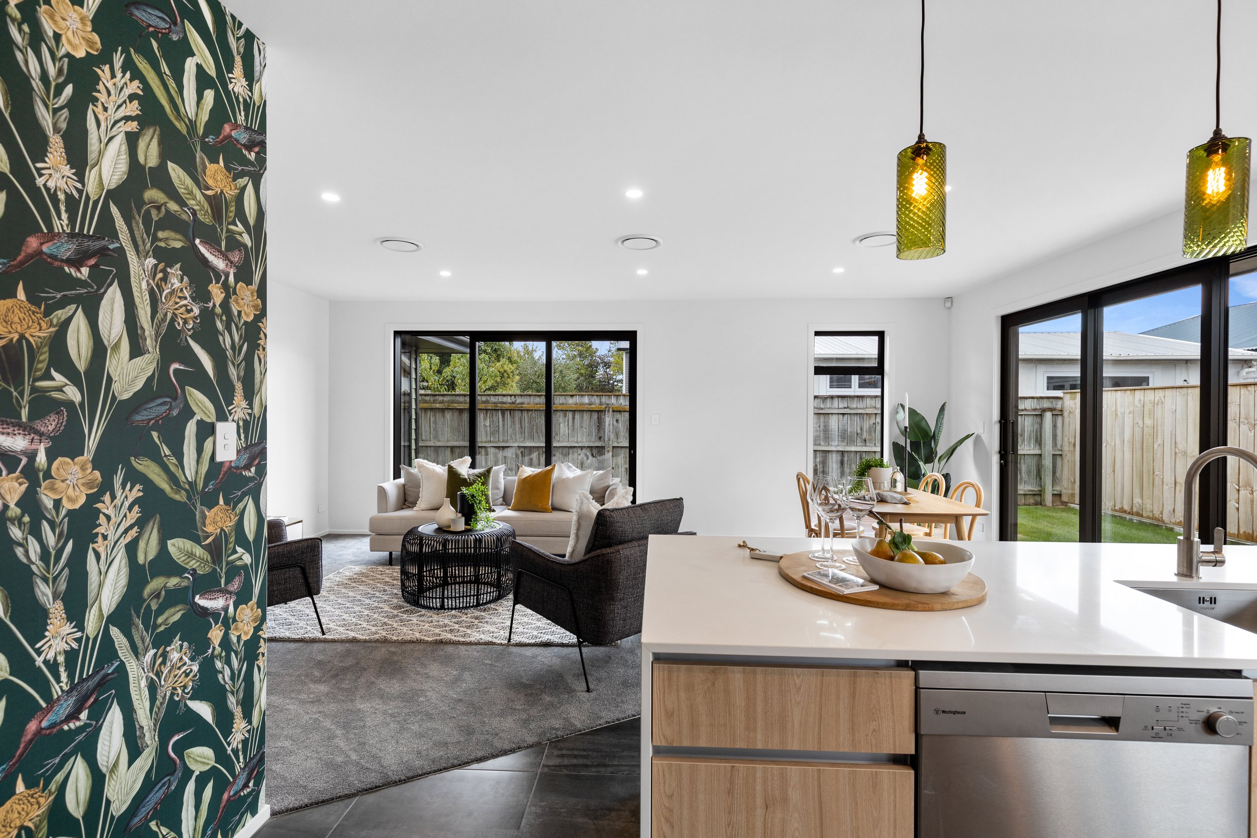 Open-concept living room and kitchen in a modern home with sliding glass doors, white walls, and hanging green pendant lights.