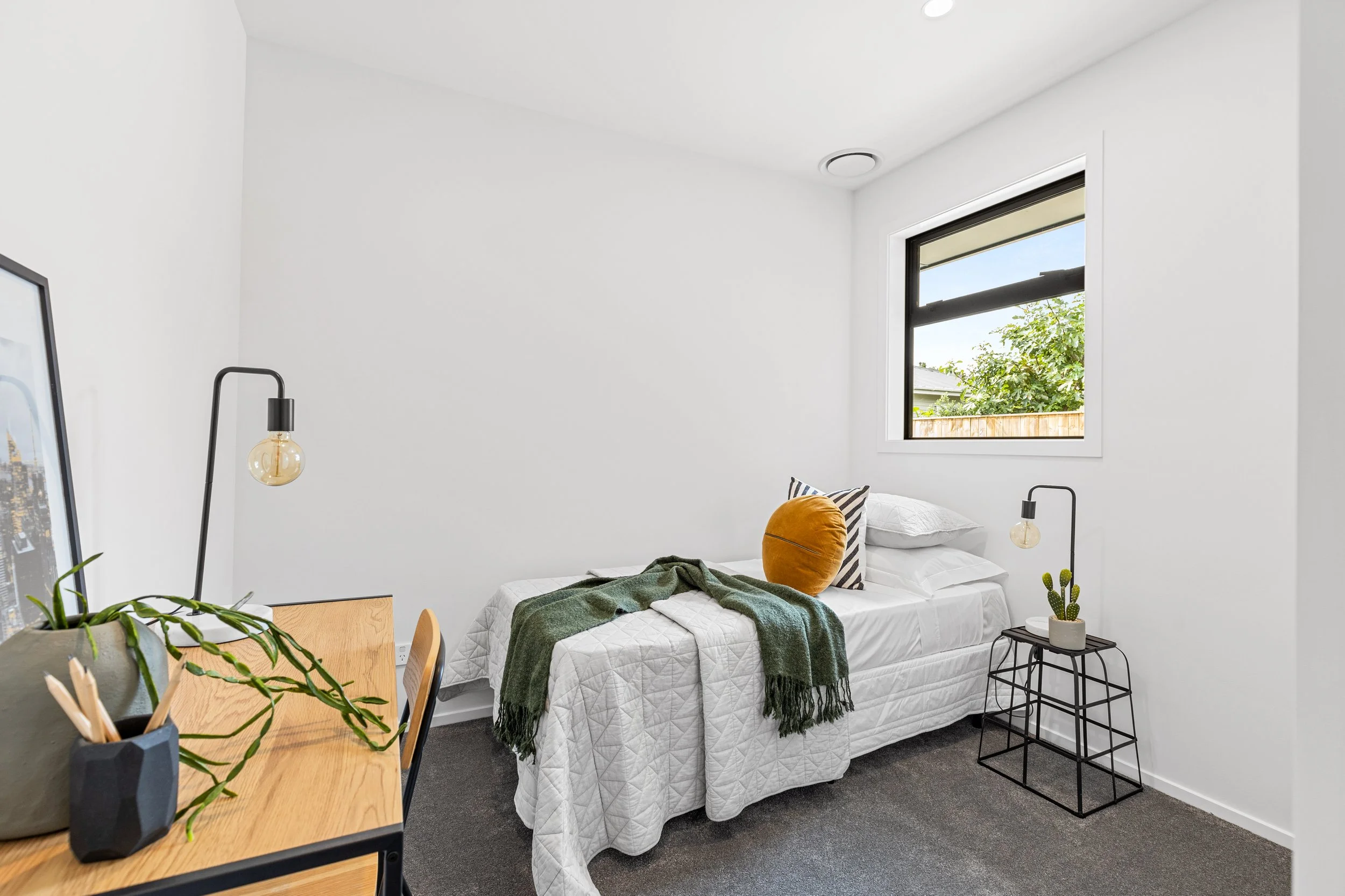 A small bedroom with a single bed covered with white bedding, a green blanket, and decorative pillows in striped, white, and orange. Next to the bed is a black metal side table with a potted cactus. The room has a window showing a tree and fence outs
