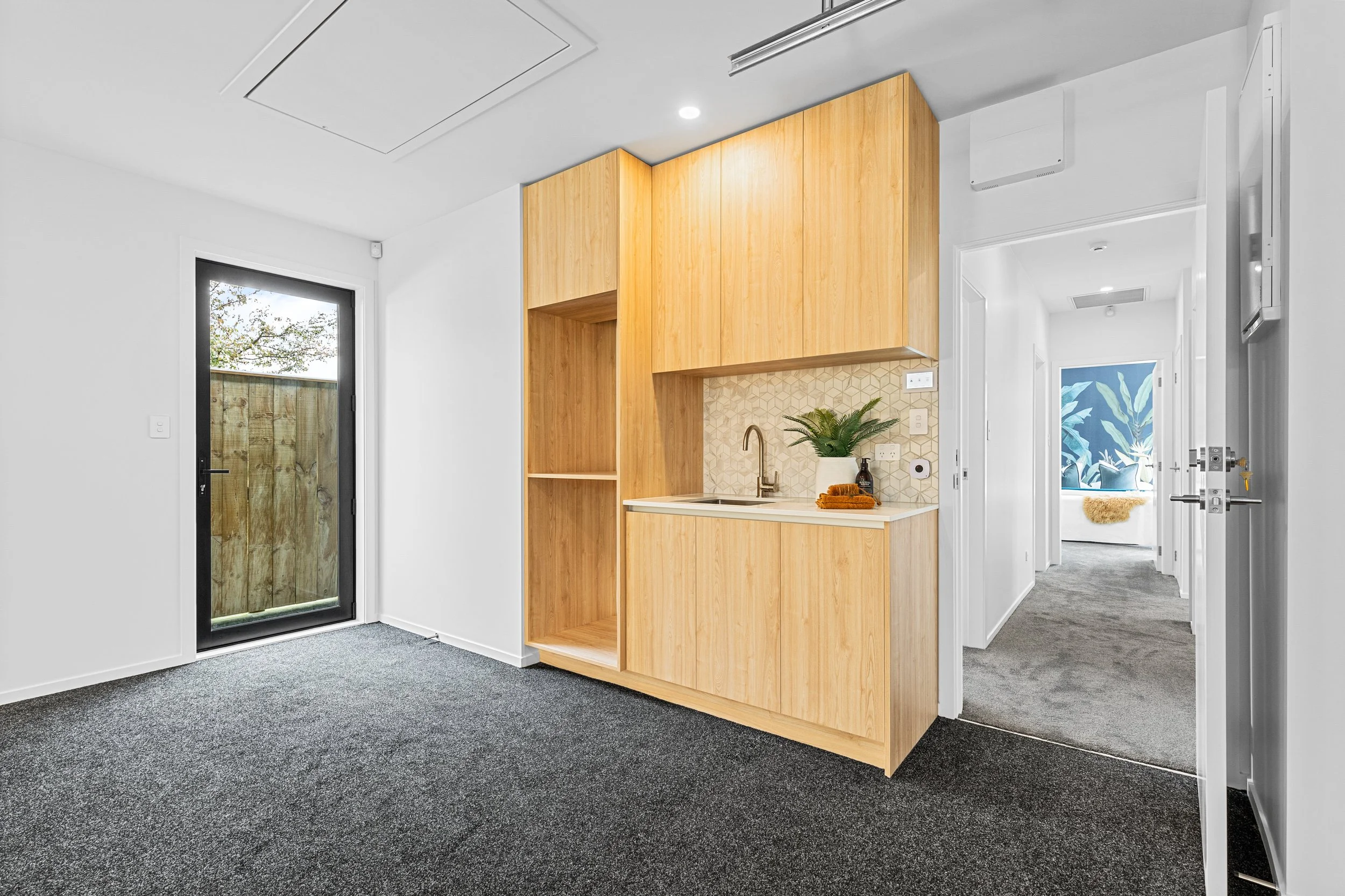 Interior of a modern apartment with a small kitchenette featuring light wood cabinets, a beige backsplash, a faucet, and a white countertop. On the counter is a potted plant and a small decorative item. To the left, there is a glass door leading outs