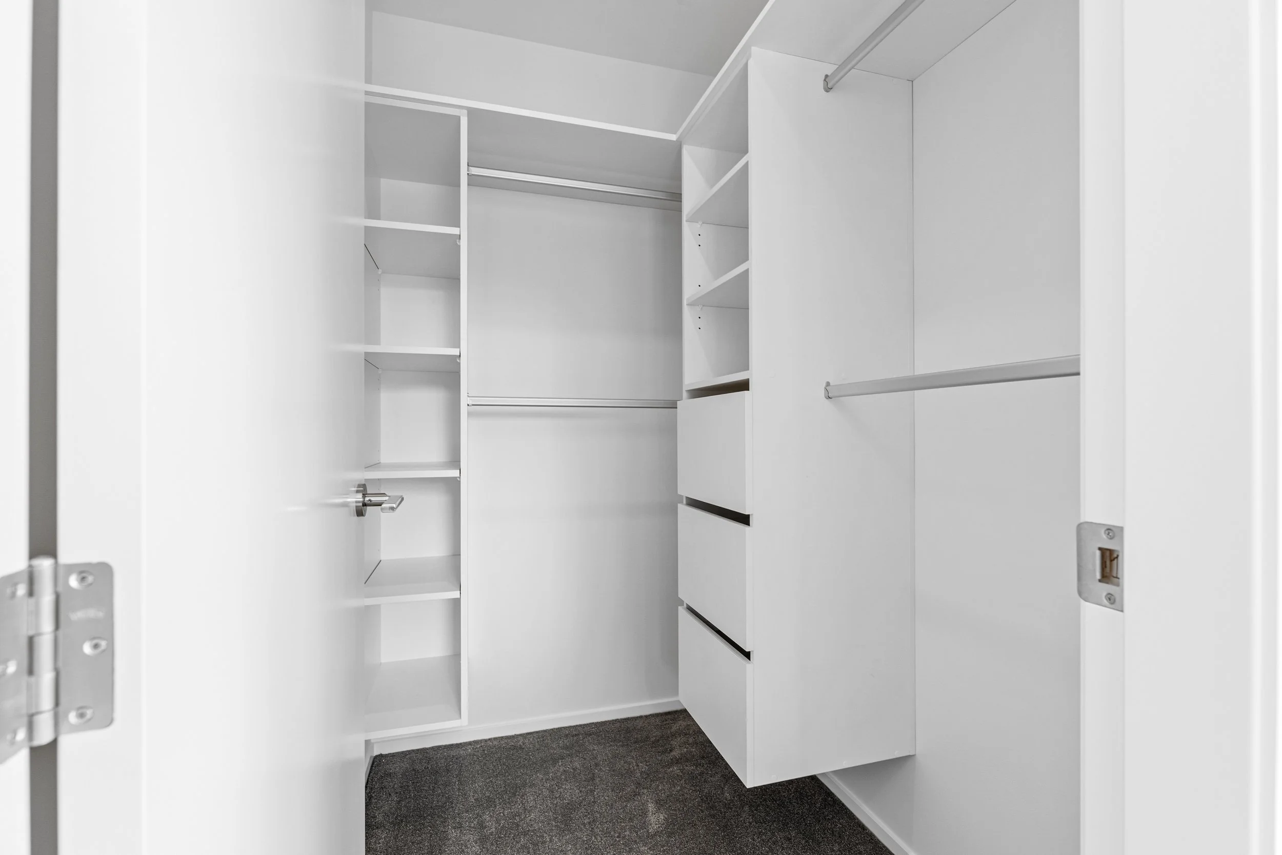 White walk-in closet with shelves and hanging rods on the walls and dark carpeted floor.