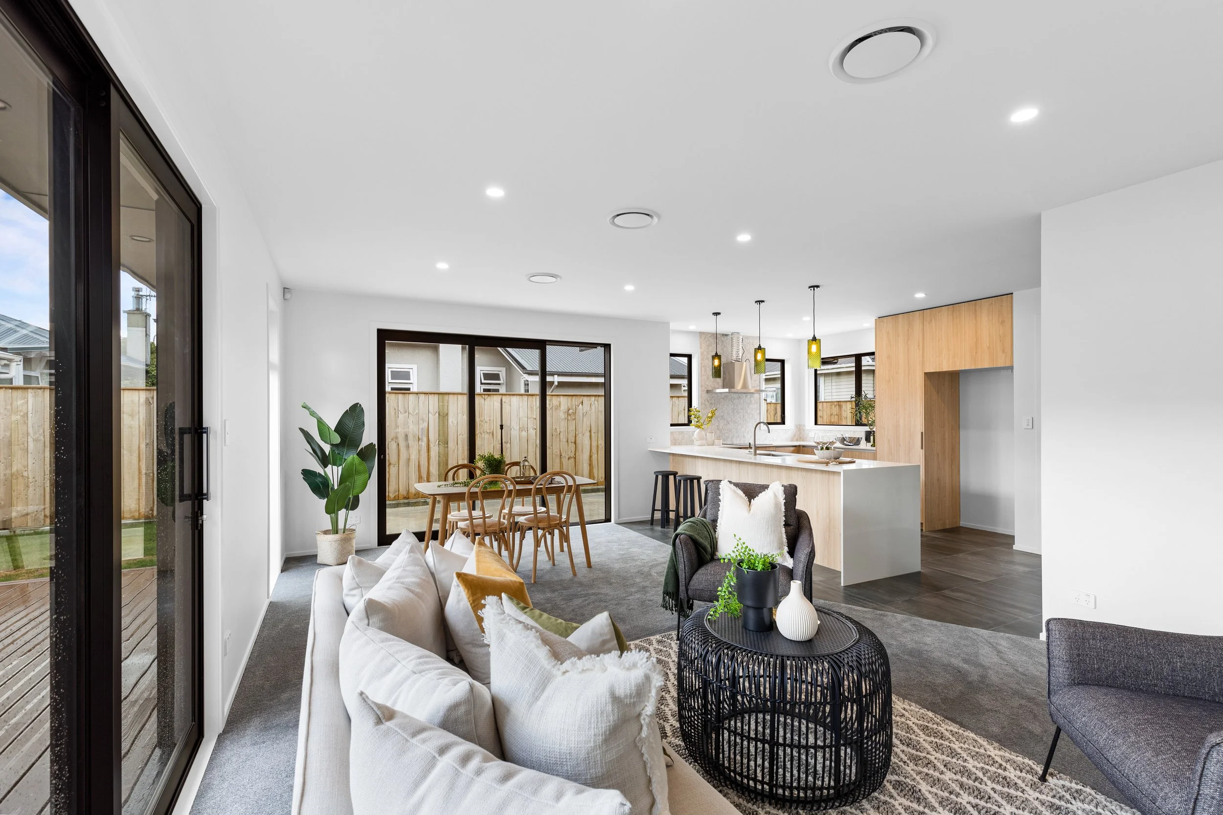 Open-concept living and dining area with white walls, grey carpet, beige sofa with white and mustard pillows, black round coffee table with decorative vases, dark grey armchair, large sliding glass doors leading to a backyard with wooden fence, woode