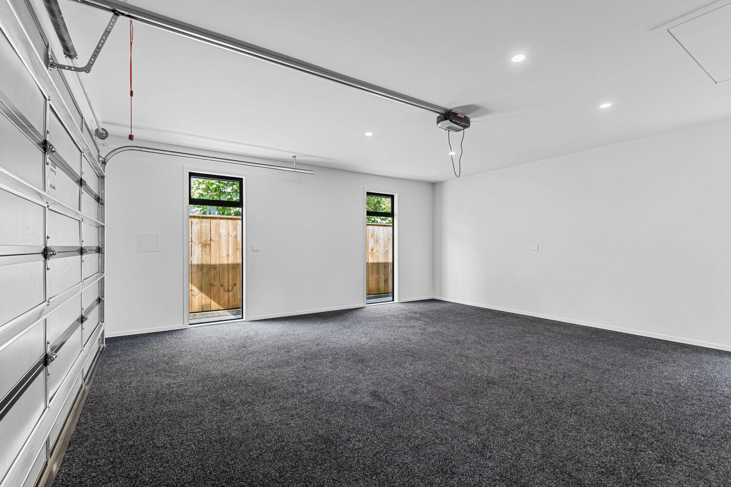 Garage with white walls, two windows, dark carpeted floor, and an open garage door with a red control cord hanging.