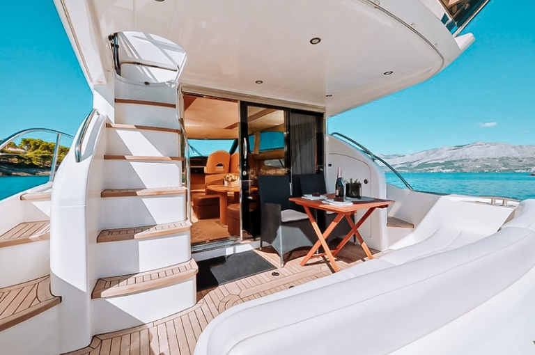 Yacht deck with outdoor seating, stairs leading to upper level, sliding glass door to interior, table with drinks, and ocean view in the background.