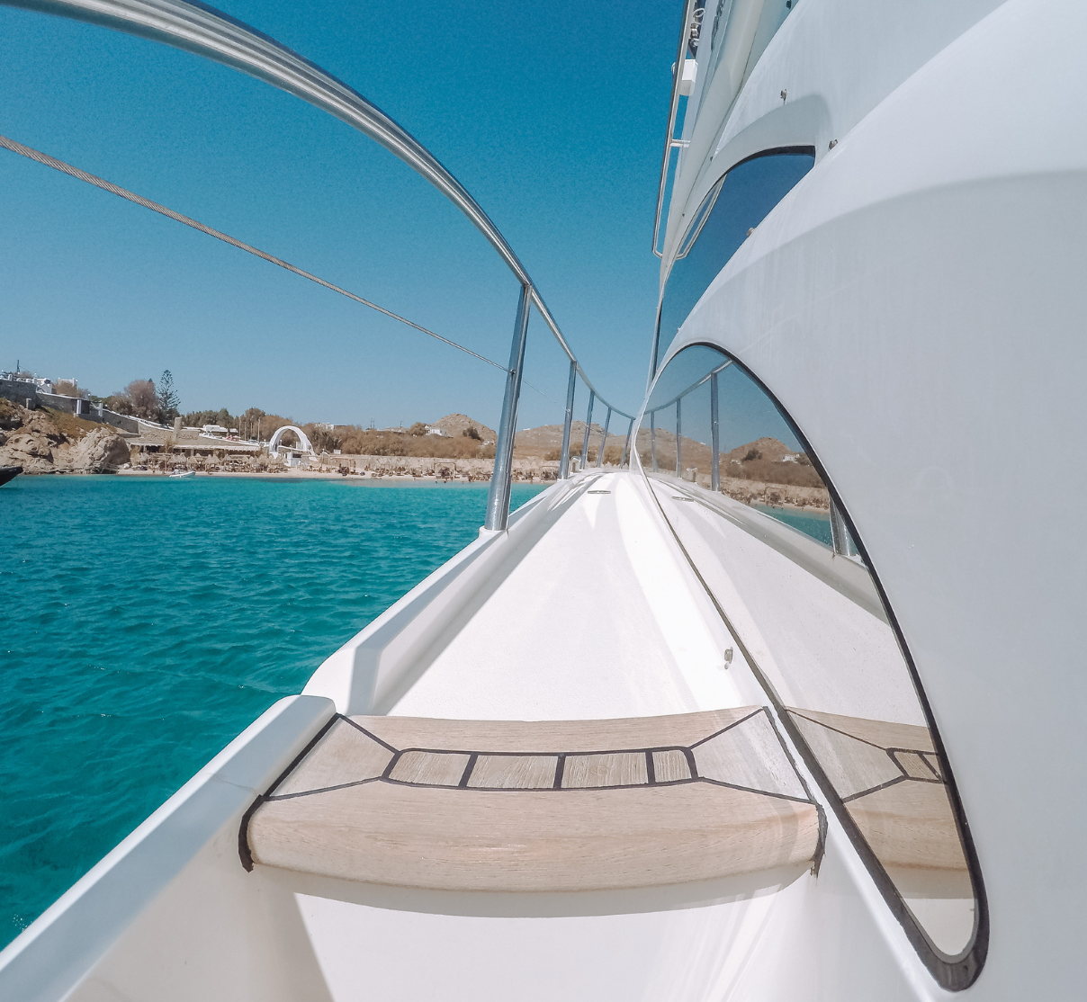 View from a yacht  in mykonos on clear turquoise water, looking toward a shoreline with brown hills and structures, under a bright blue sky.
