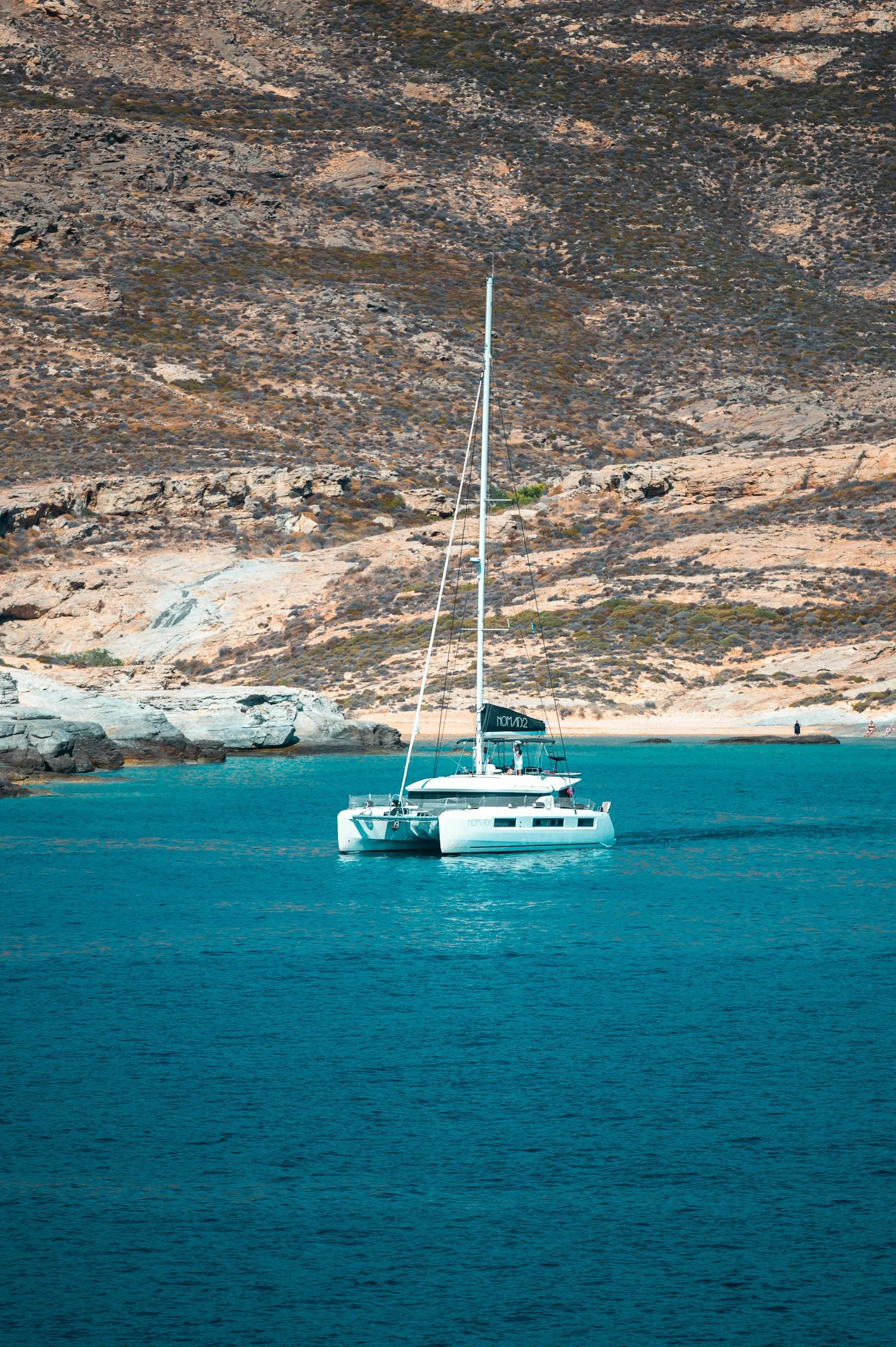 A white sailboat named 'NOVEL' floating on clear blue water near a rocky shoreline with brown and gray hills in the background.