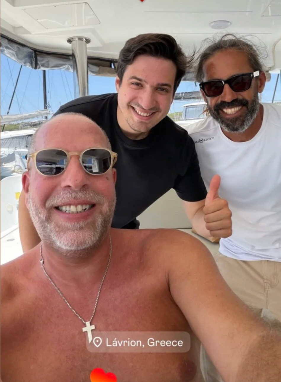 Three men taking a selfie aboard a boat in Lávion, Greece. The man in the foreground is shirtless, wearing sunglasses, a necklace with a cross, and smiling. The other two men are behind him, smiling, with one giving a thumbs up. The man on the left h