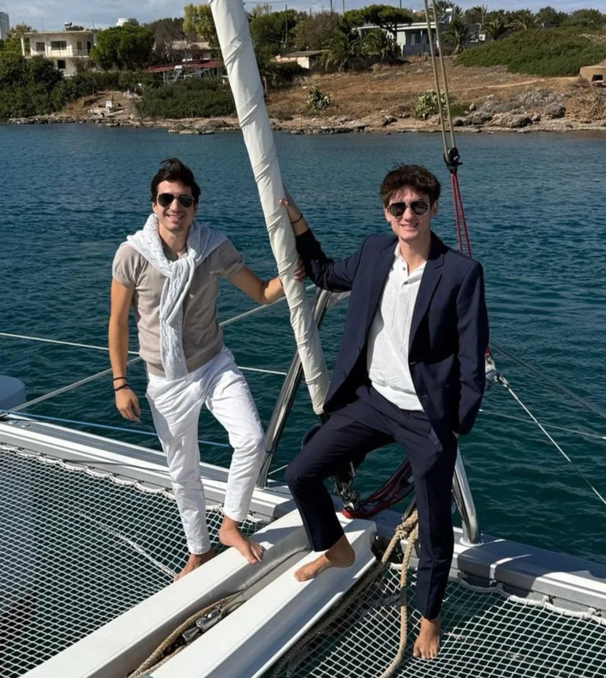 Two young men in sunglasses on a sailboat, smiling. One is dressed casually, the other in a suit, both barefoot. They are near a shoreline with houses and greenery.