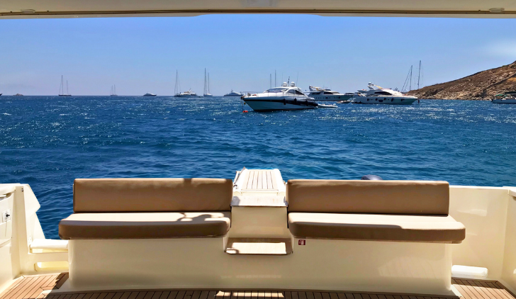 View from a yacht showing calm blue water with several other yachts anchored, a hilly coastline, and a clear blue sky.