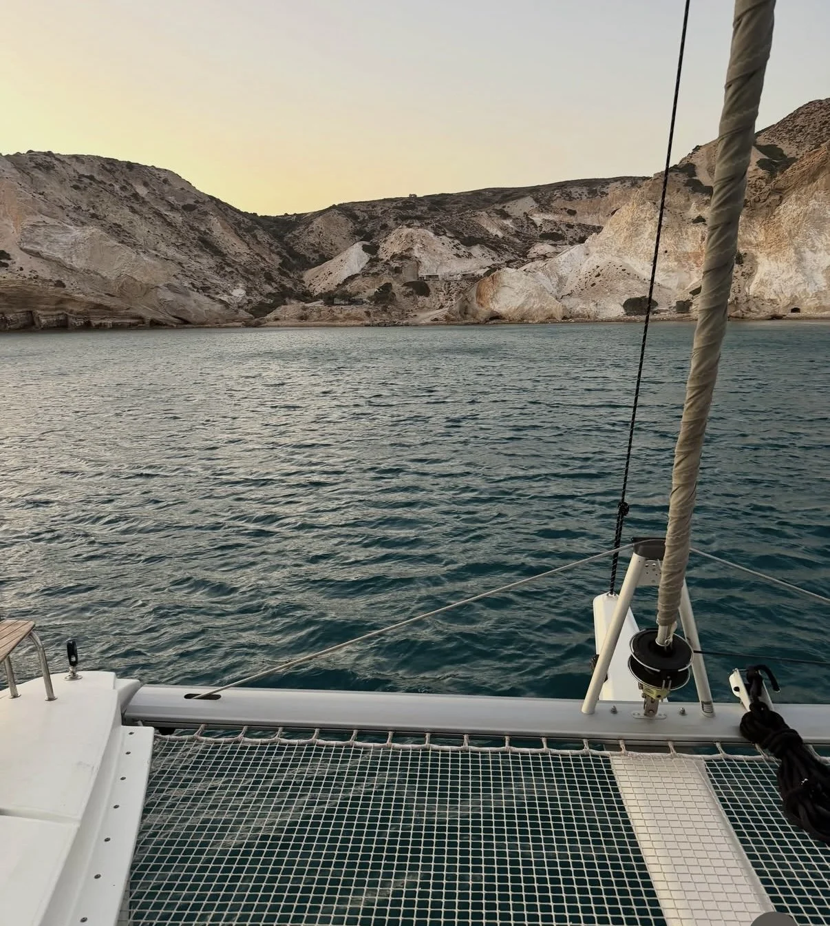 View from a boat showing calm waters, rocky hills in the background, and part of the boat's deck with ropes, a seating area, and a support pole.