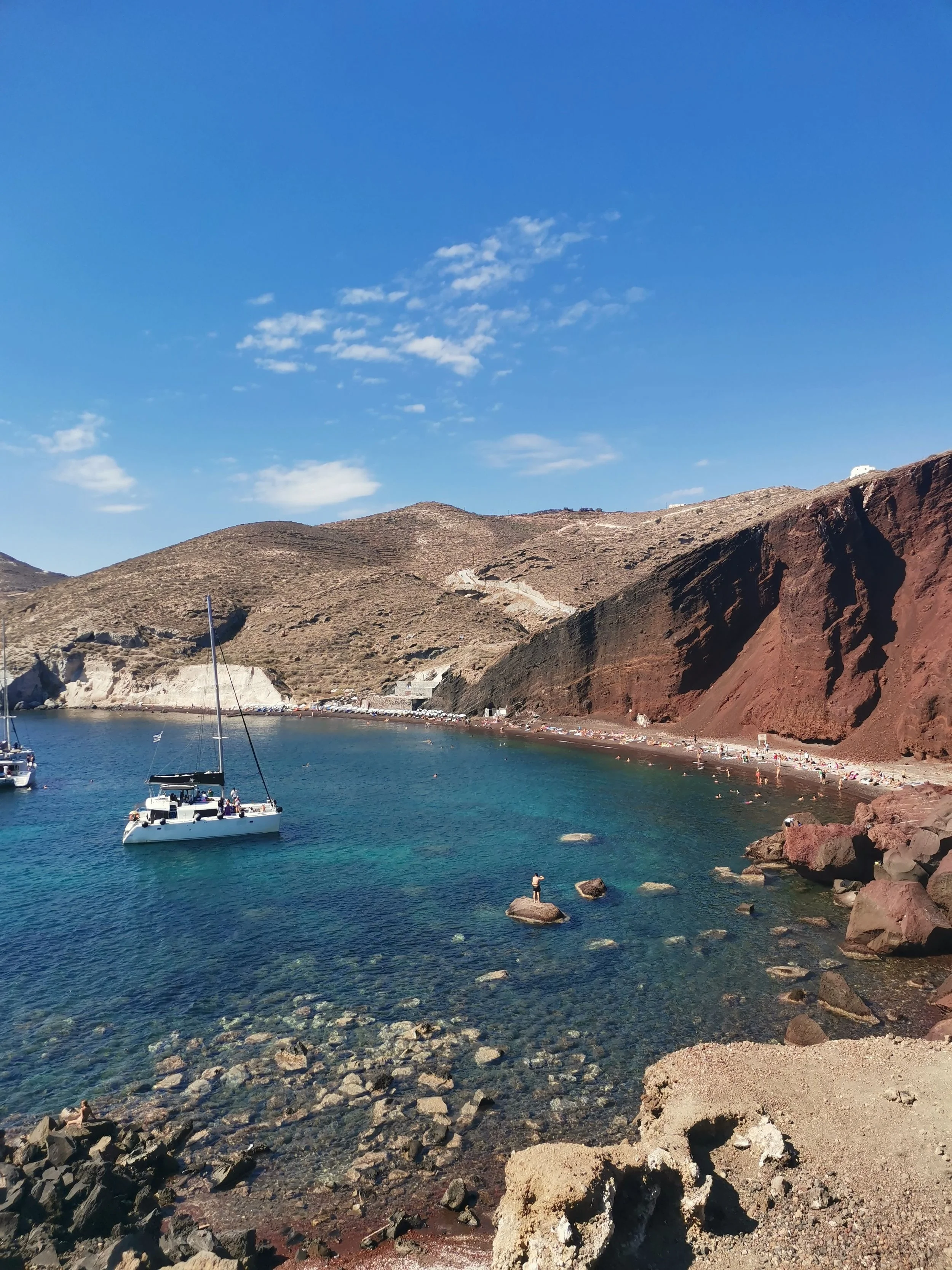 A coastal scene with boats anchored near a rocky shore. Brown hills with sparse vegetation rise in the background under a clear blue sky with some clouds.