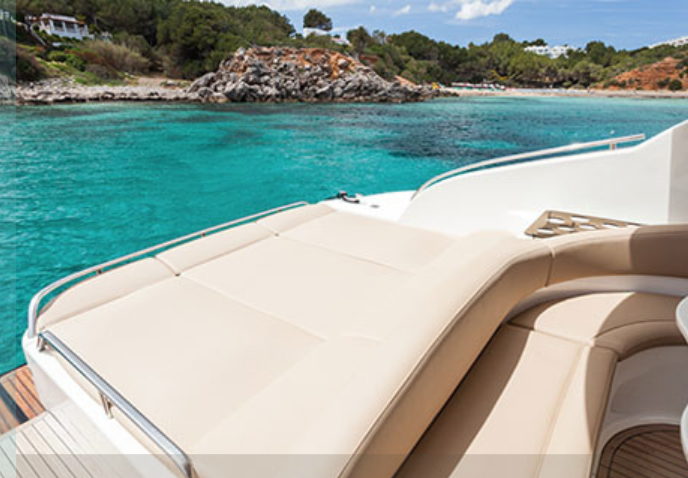 View of a yacht with beige seating on a turquoise ocean with a rocky shoreline and green trees in the background.