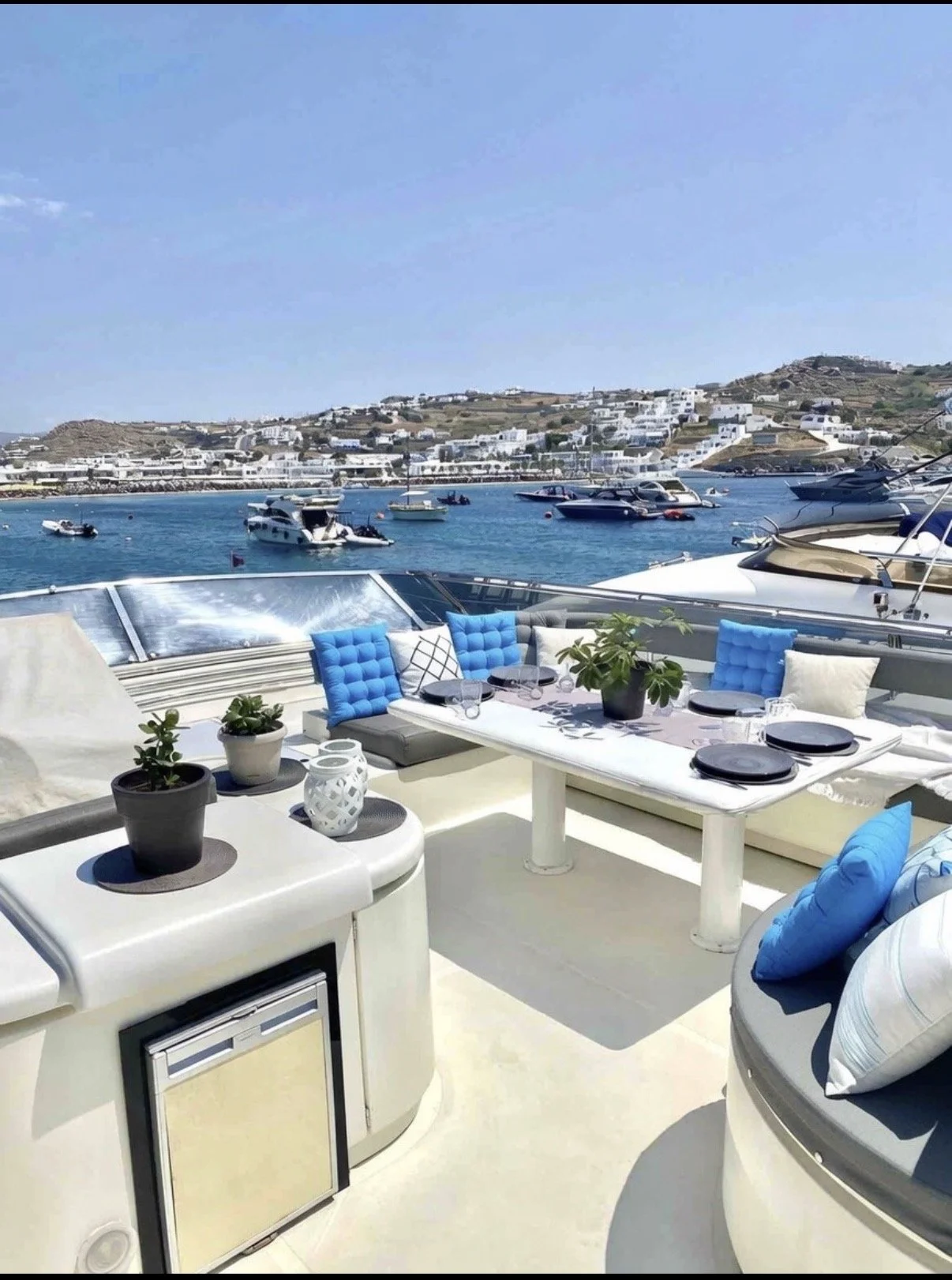 View of a yacht deck with blue cushions and potted plants, overlooking the ocean with numerous boats and white buildings on hillside in the background during daytime.