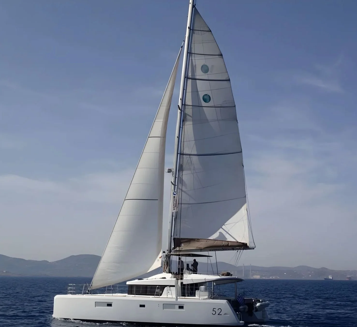 A large white sailing yacht with multiple sails on open water, with two people on deck and distant land in the background under a clear blue sky.