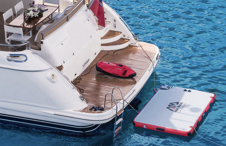 Back of a yacht with a swimming ladder, a red paddleboard, and a floating platform with the M4 logo, surrounded by blue water.