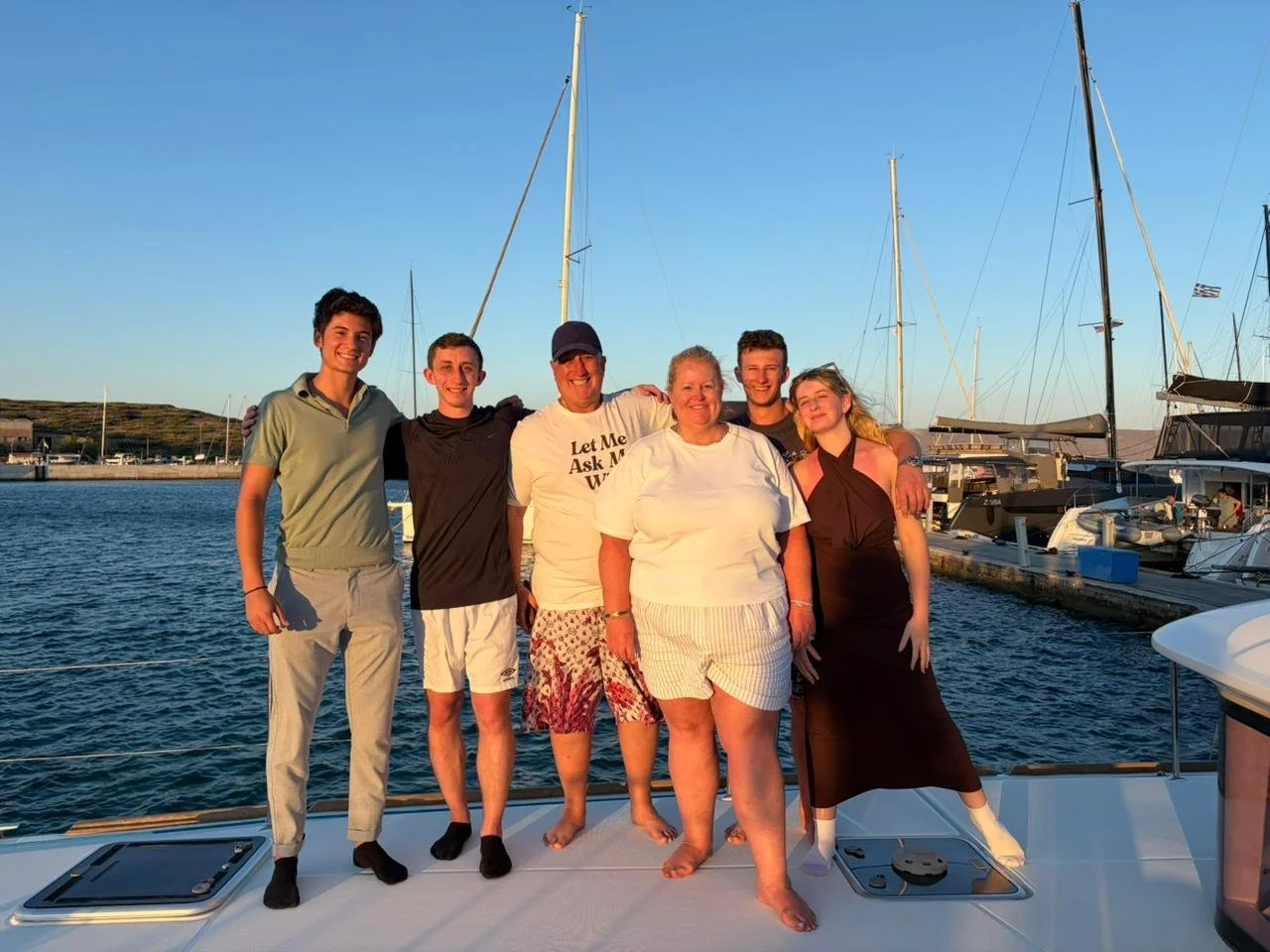 A group of six people standing together on a boat docked at a marina during sunset, with sailboats in the background.