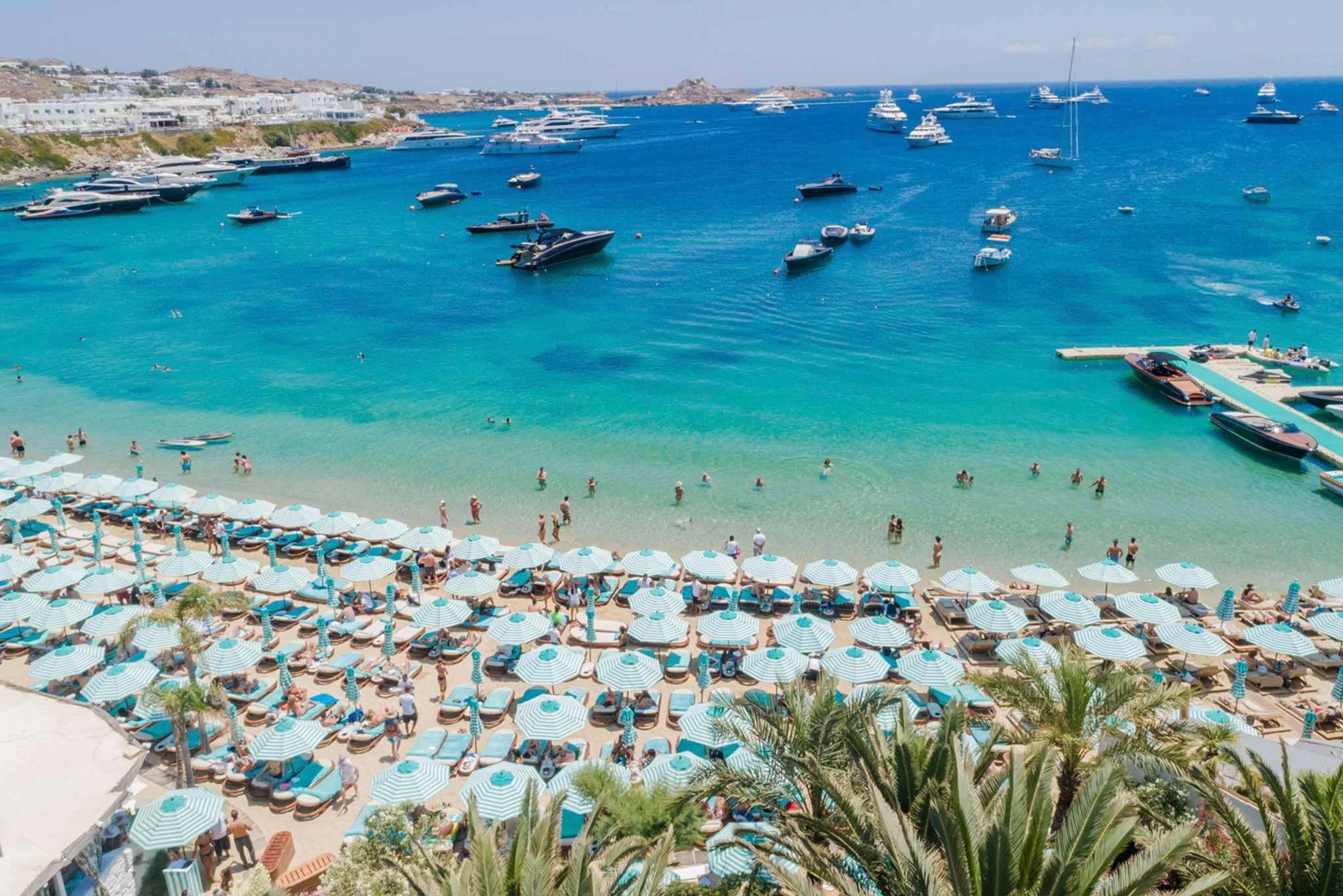Beach with white and blue striped umbrellas, people swimming, and yachts anchored in turquoise water, hillside with buildings in the background.