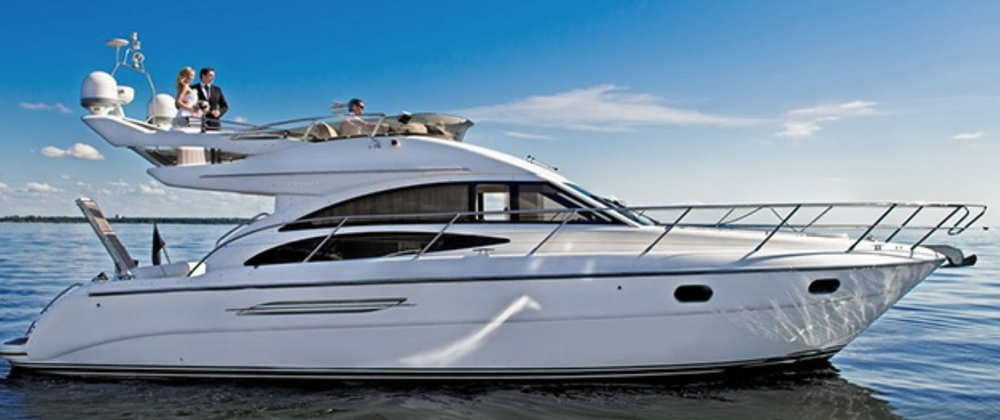 A white yacht with multiple decks on calm water under a blue sky with some clouds, with four people on the top deck.