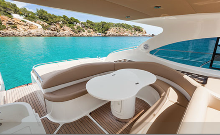 Luxury yacht deck with curved seating area and table overlooking turquoise sea and rocky coastline with green trees.