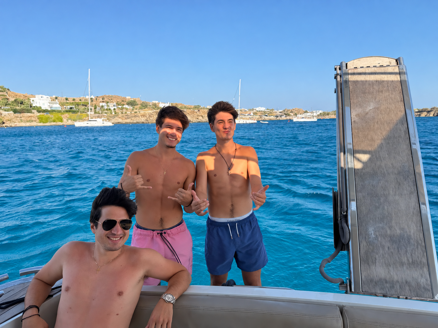 Three young men on a boat in the water with a coastal landscape in the background. Two are standing, shirtless, making hand gestures. The third, sitting, is wearing sunglasses and a watch, smiling.