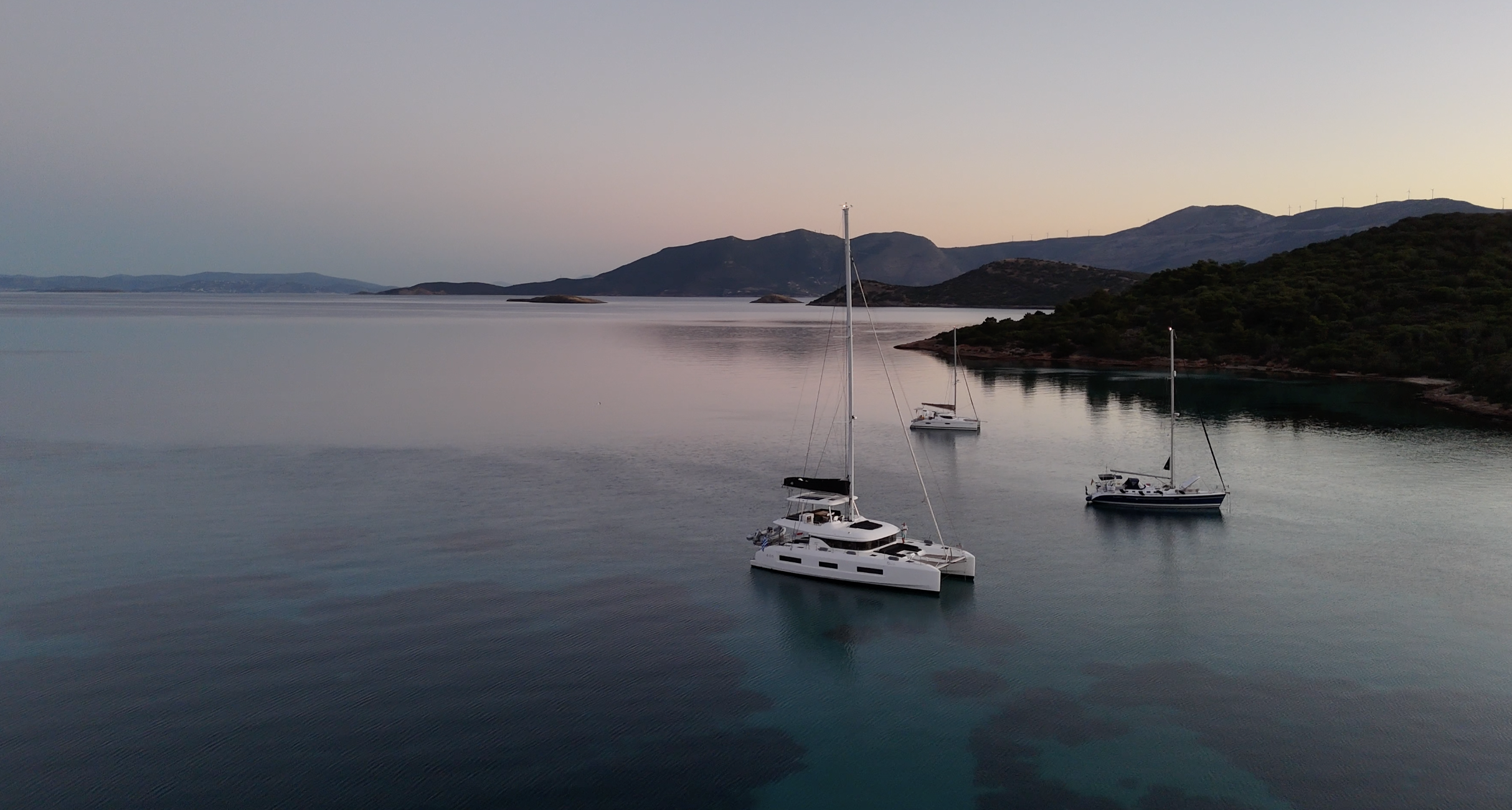Three sailboats anchored in calm water near a forested shoreline with mountains in the background, under a pale sky.