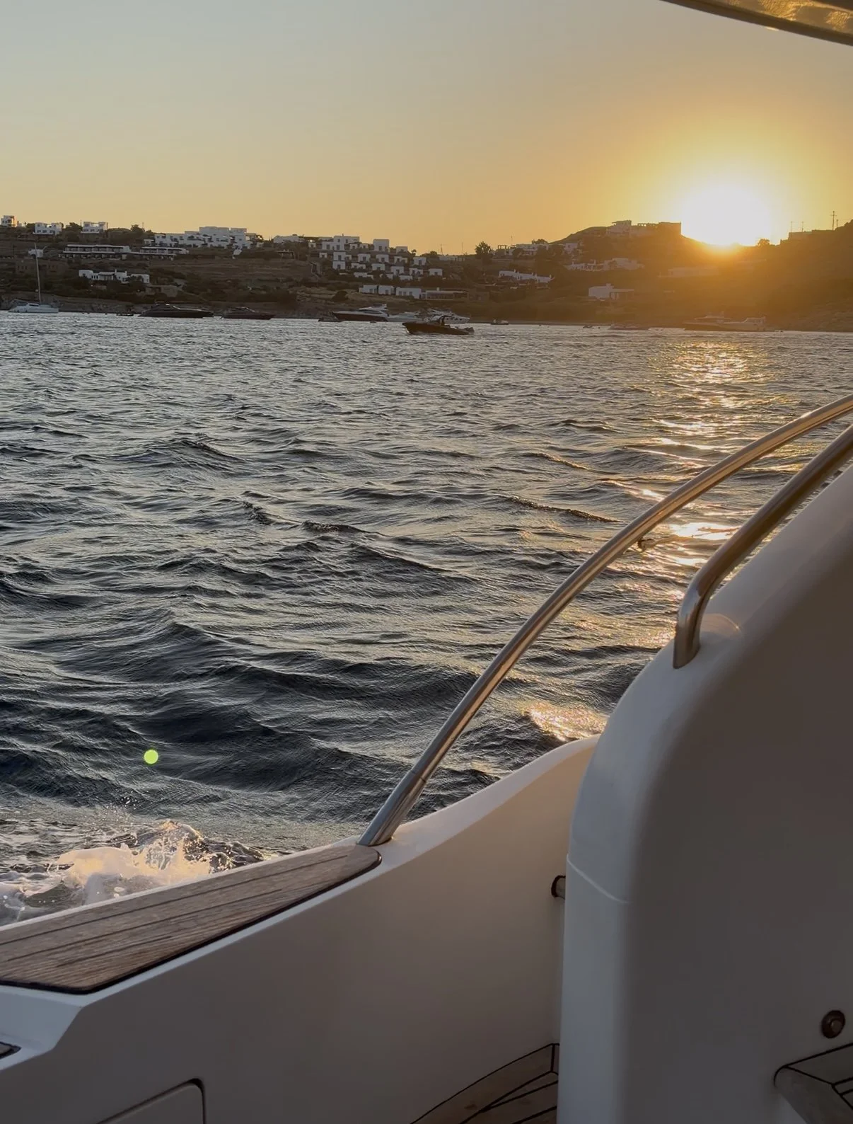 Sunset view from a boat showing water with ripples, boats in the distance, and a hillside with white buildings illuminated by the setting sun.