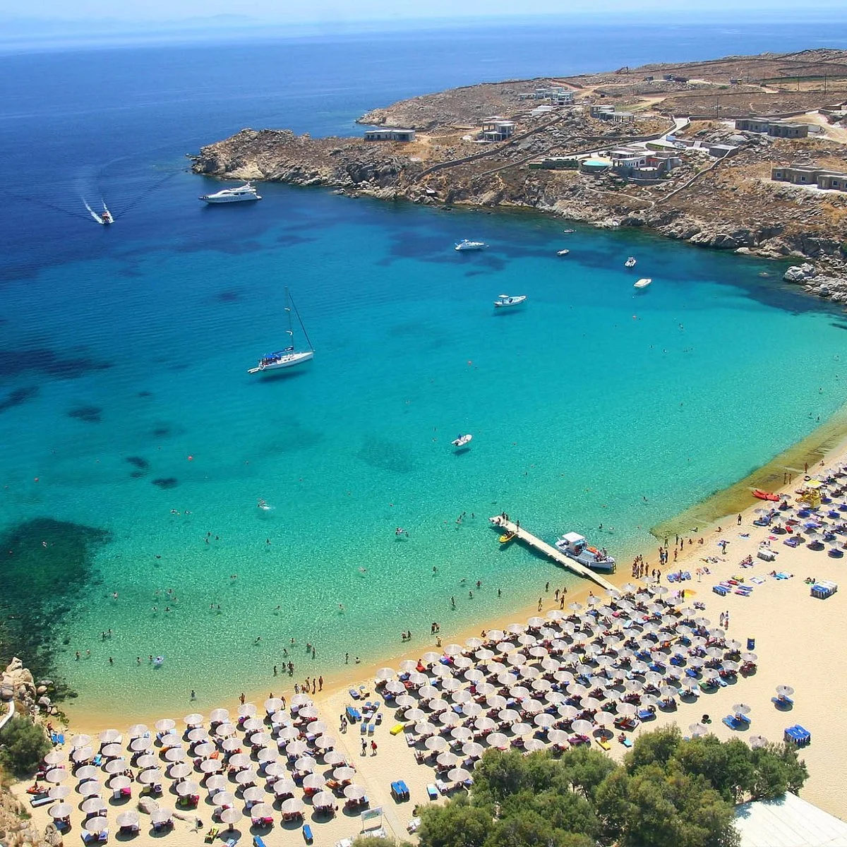A beach with numerous umbrellas, sunbathers, and swimmers, turquoise water with boats, and rocky cliffs in the background.