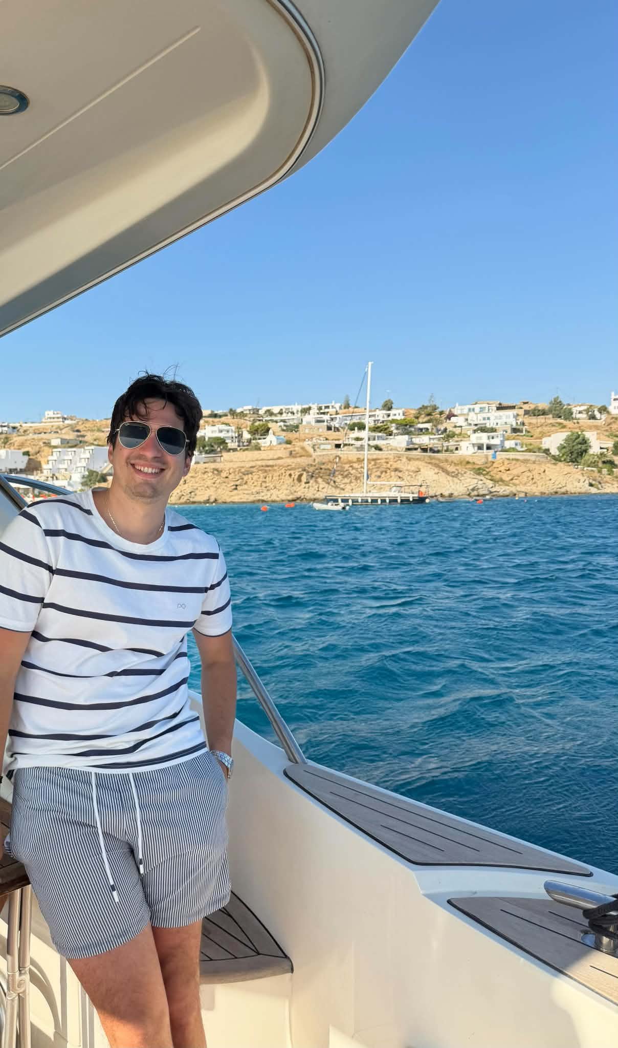 A man smiling in sunglasses standing on a boat, with water and a hillside with houses in the background.
