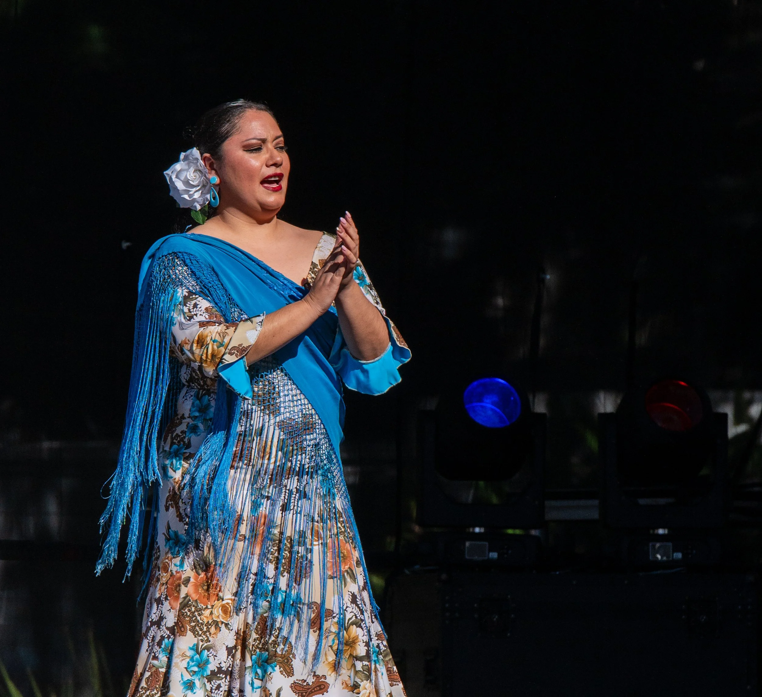 A woman wearing traditional Mexican dress with floral and butterfly patterns, a vibrant blue shawl, and floral hair accessory performs a dance or song on stage with dark background and stage lights.