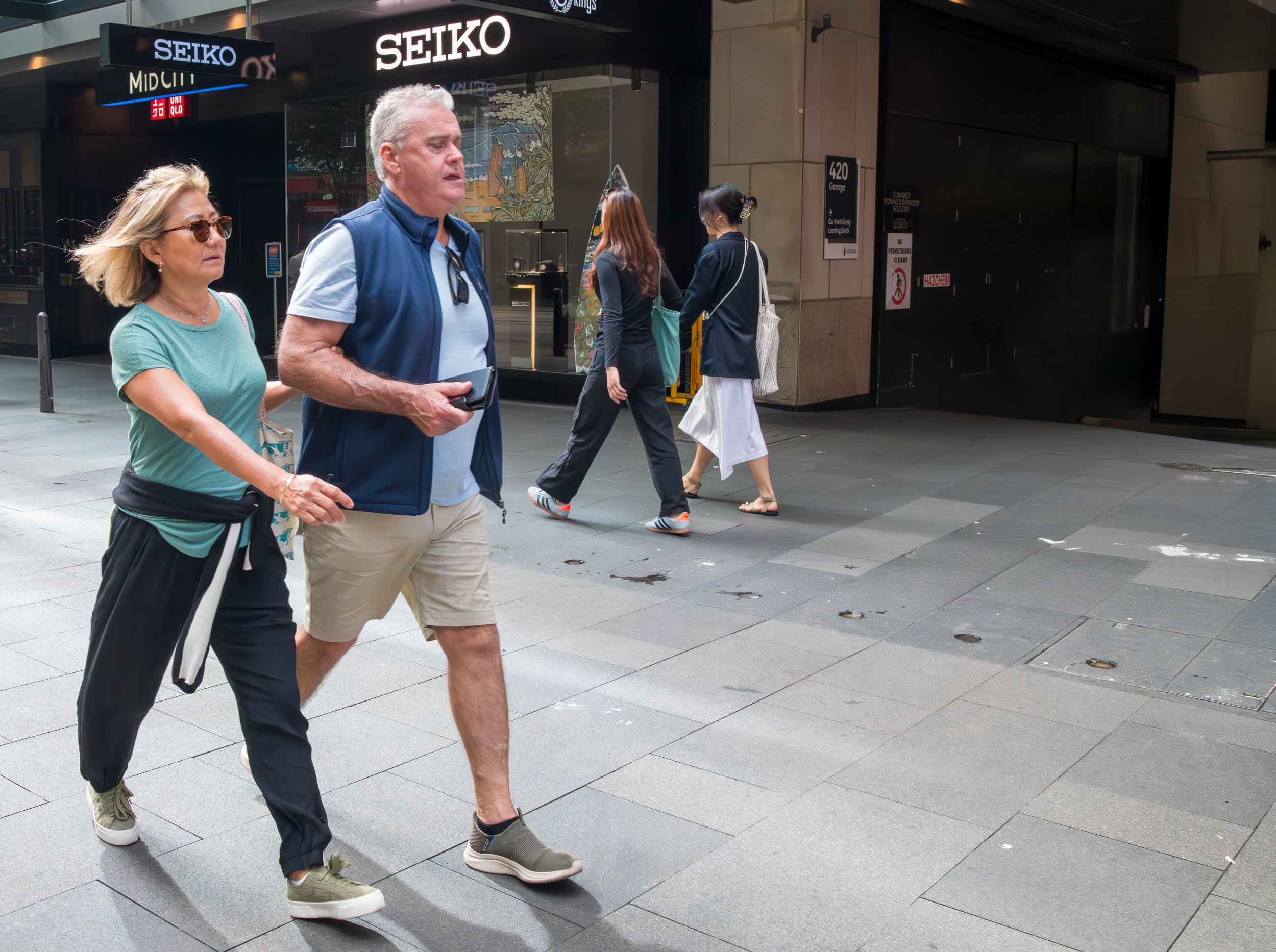 Three people walking on a city sidewalk near a Seiko store with two women and one man in front, and two women walking behind them, all dressed in casual clothing.