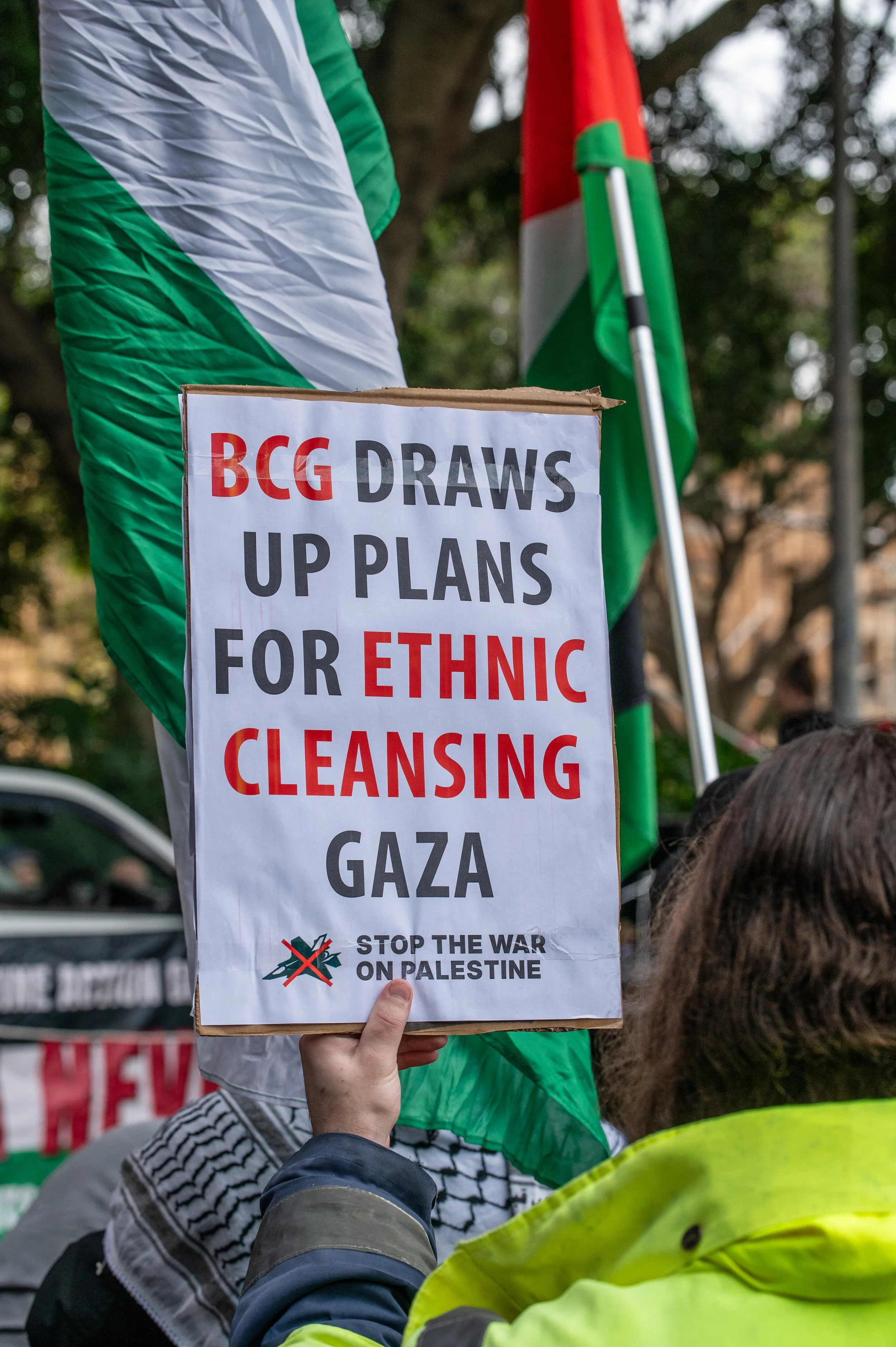 A protest sign held up among a crowd, reading: "BCG DRAWS UP PLANS FOR ETHNIC CLEANSING GAZA," with a crossed-out Palestinian flag and the message "STOP THE WAR ON PALESTINE."