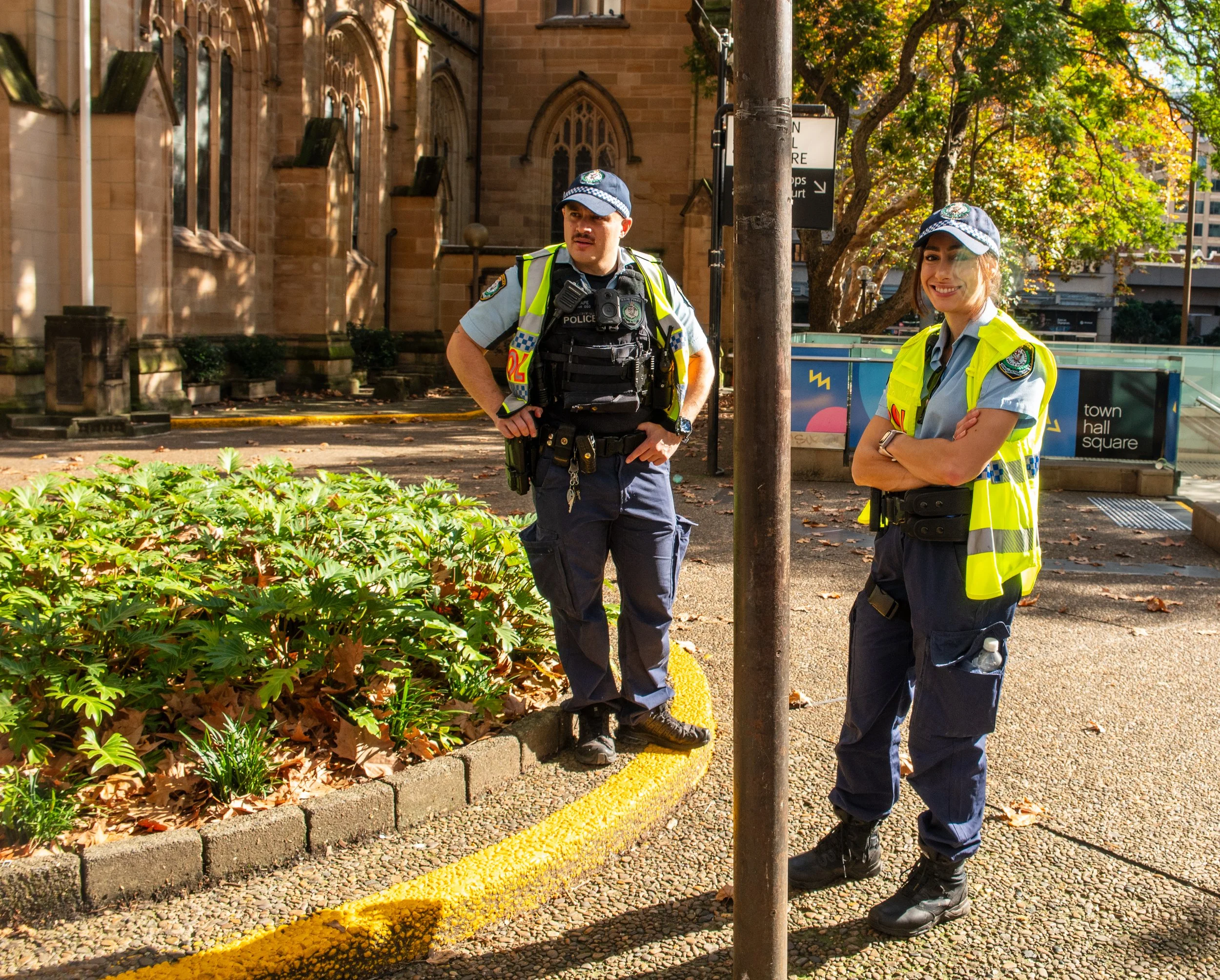 Two police officers standing on a sidewalk near a garden bed with green plants and fallen leaves, in front of a brick building with arched windows, during daytime with sunlight and trees in the background.