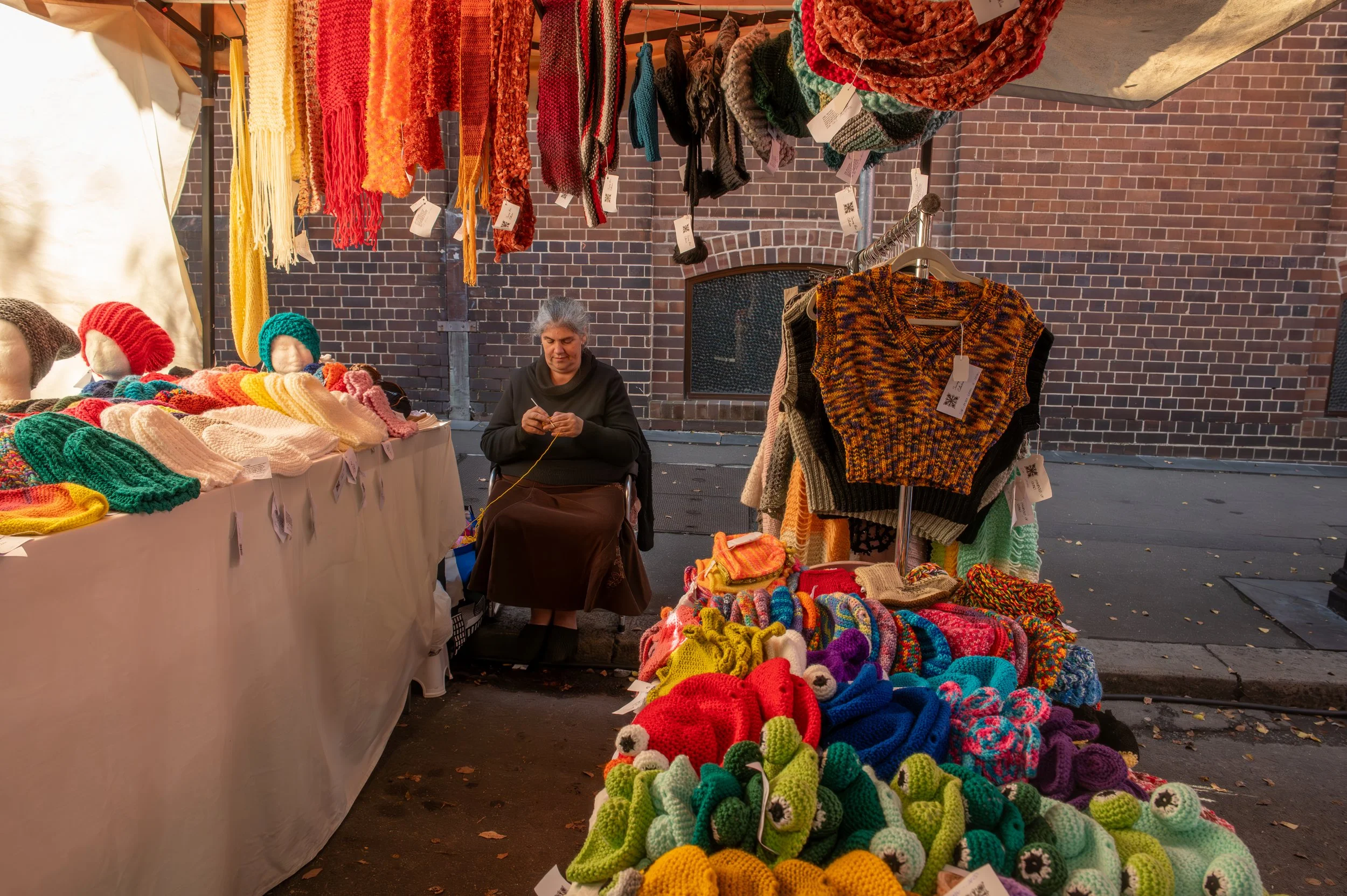 A woman sitting at a street vendor stall with colorful handmade crocheted hats, scarves, and sweaters displayed on tables and hanging above her, set against a brick wall background.