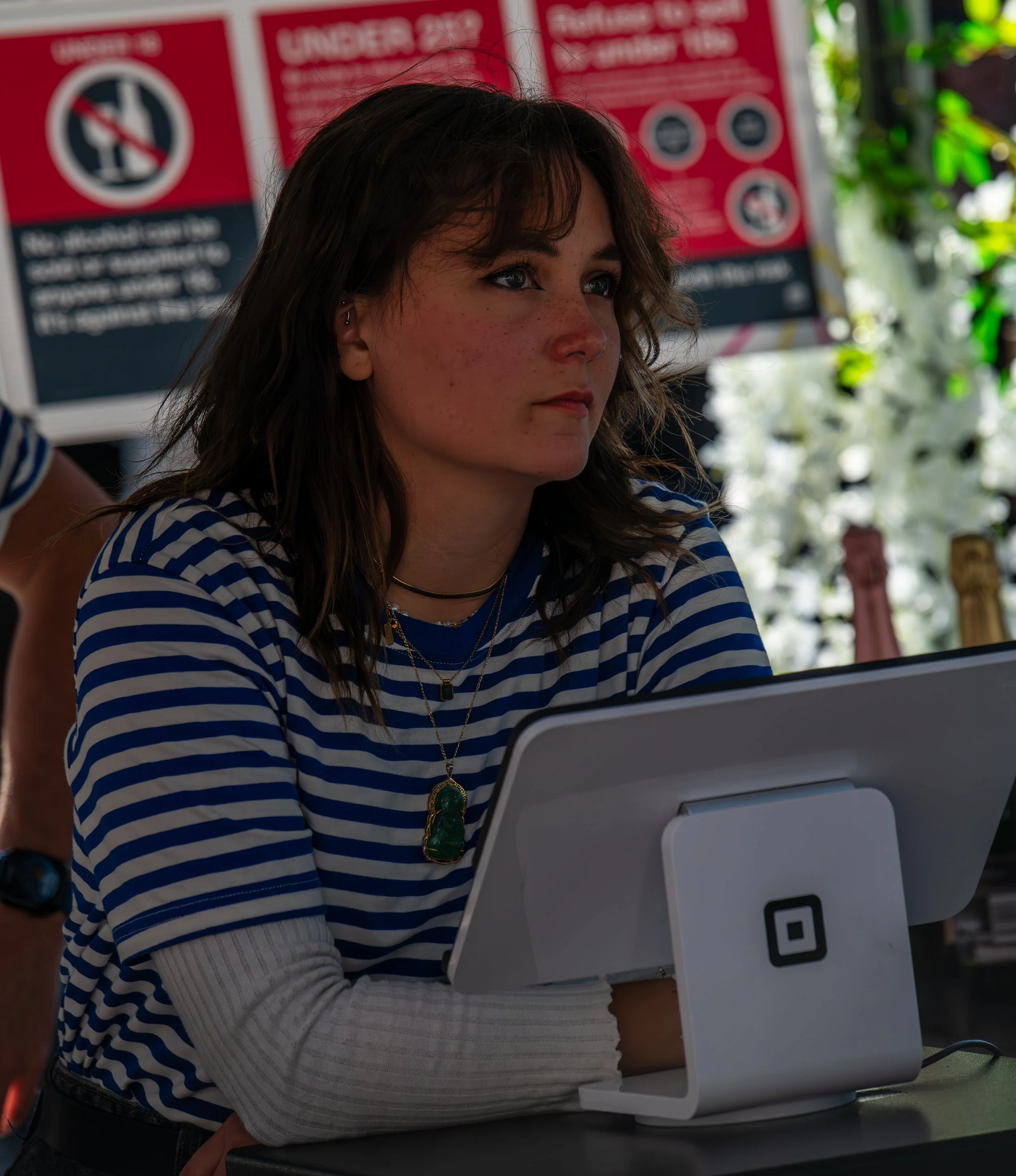 A woman with wavy brown hair, wearing a blue and white striped shirt, sitting at a table with a small white device in front of her, in an outdoor setting with red warning signs and greenery in the background.