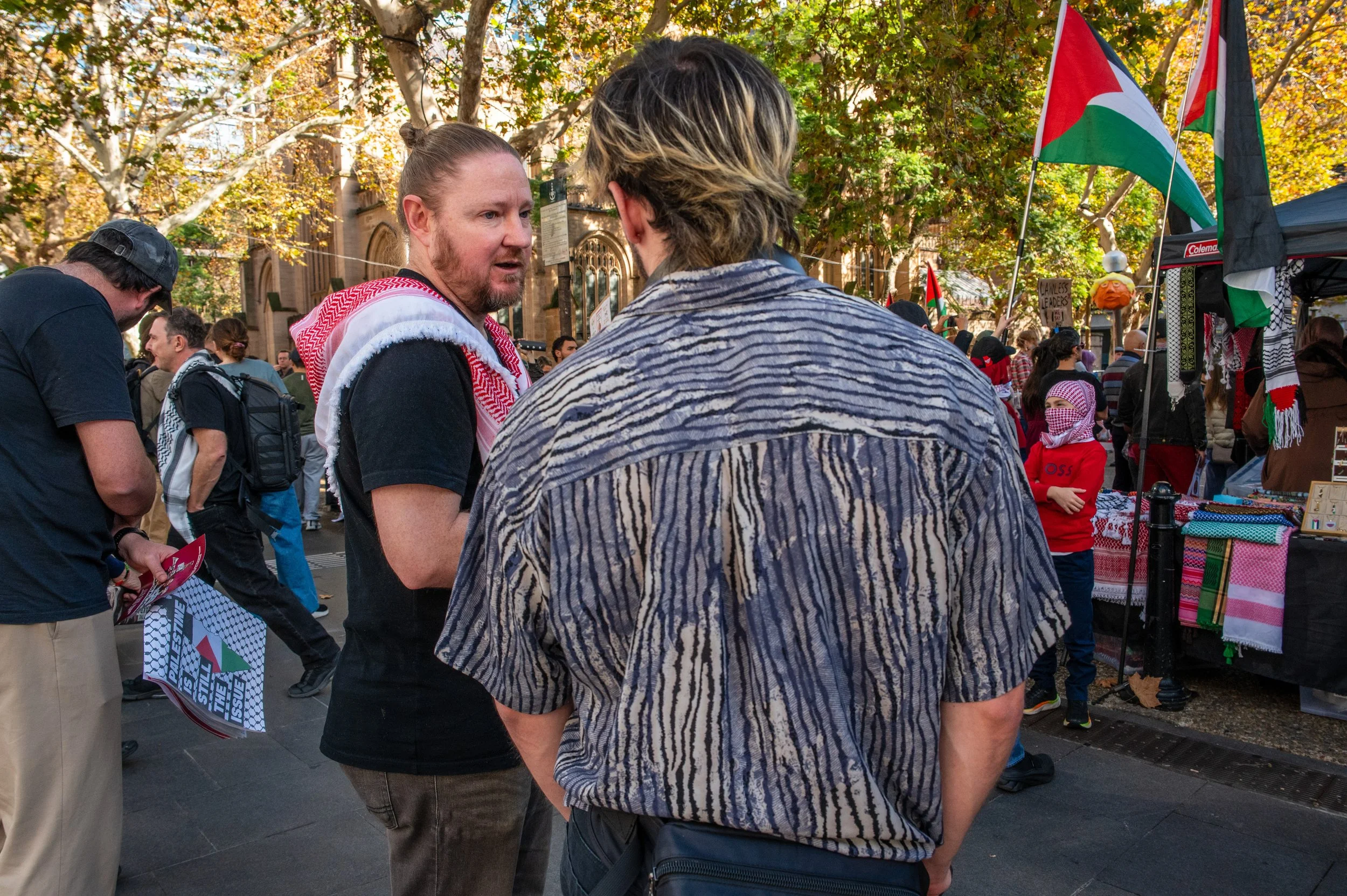 Two men engaged in conversation at a street protest or gathering, with a crowd, flags, and vendor stalls in the background.