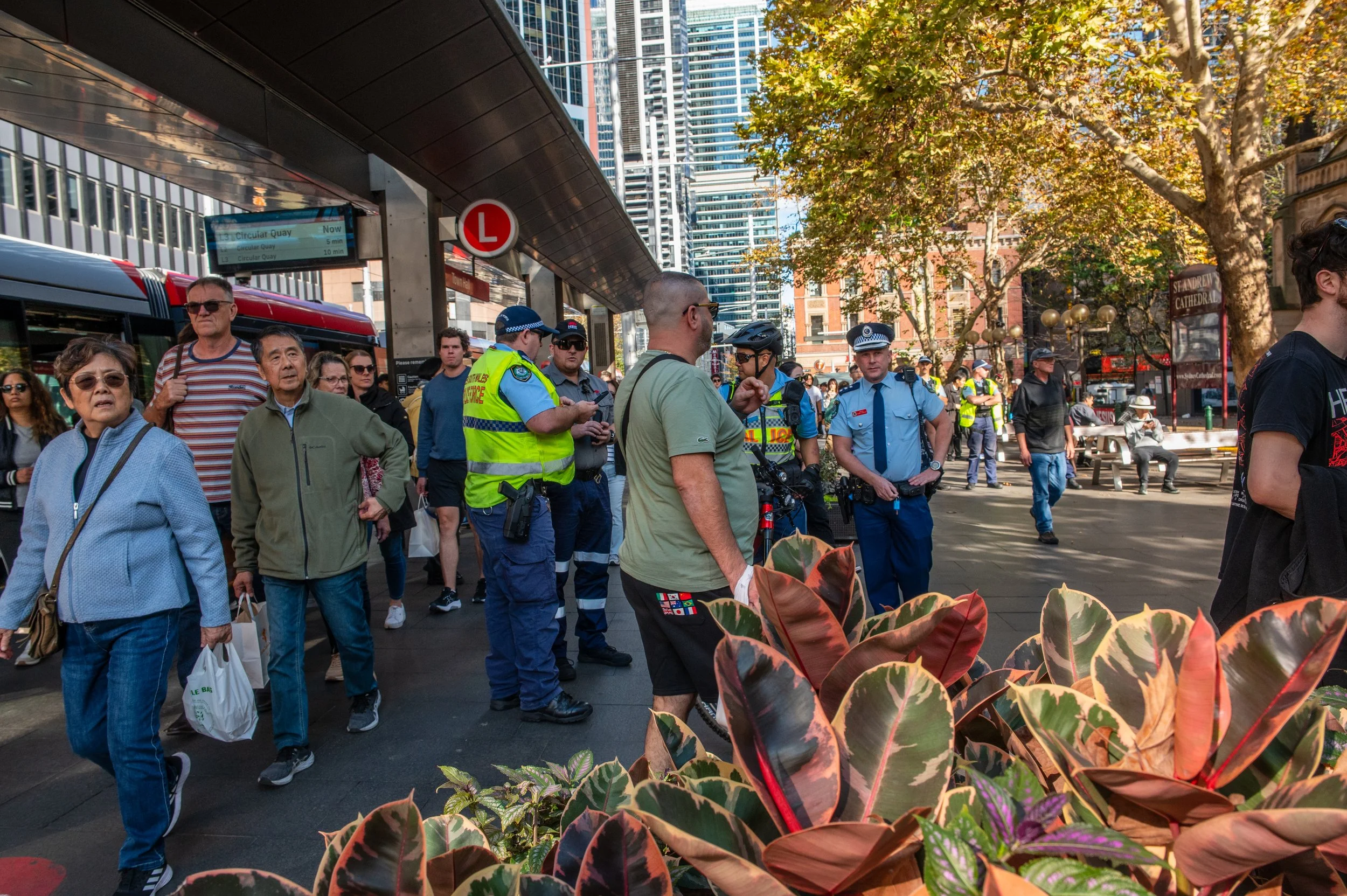 Crowd of people, including police officers and civilians, waiting at a bus stop in an urban area on a sunny day. There are tall buildings and trees in the background, with a public transportation sign and a bus nearby.