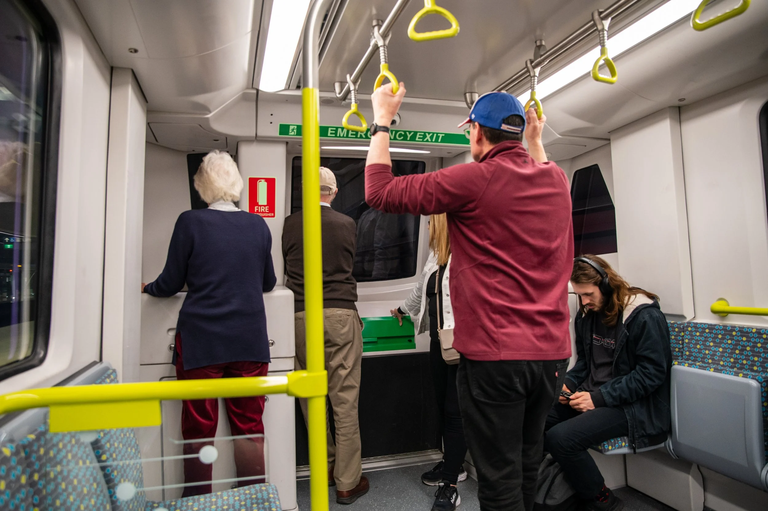 People standing and sitting in a subway train, some looking at their phones, one woman at the front near the emergency exit, others holding onto yellow handrails.