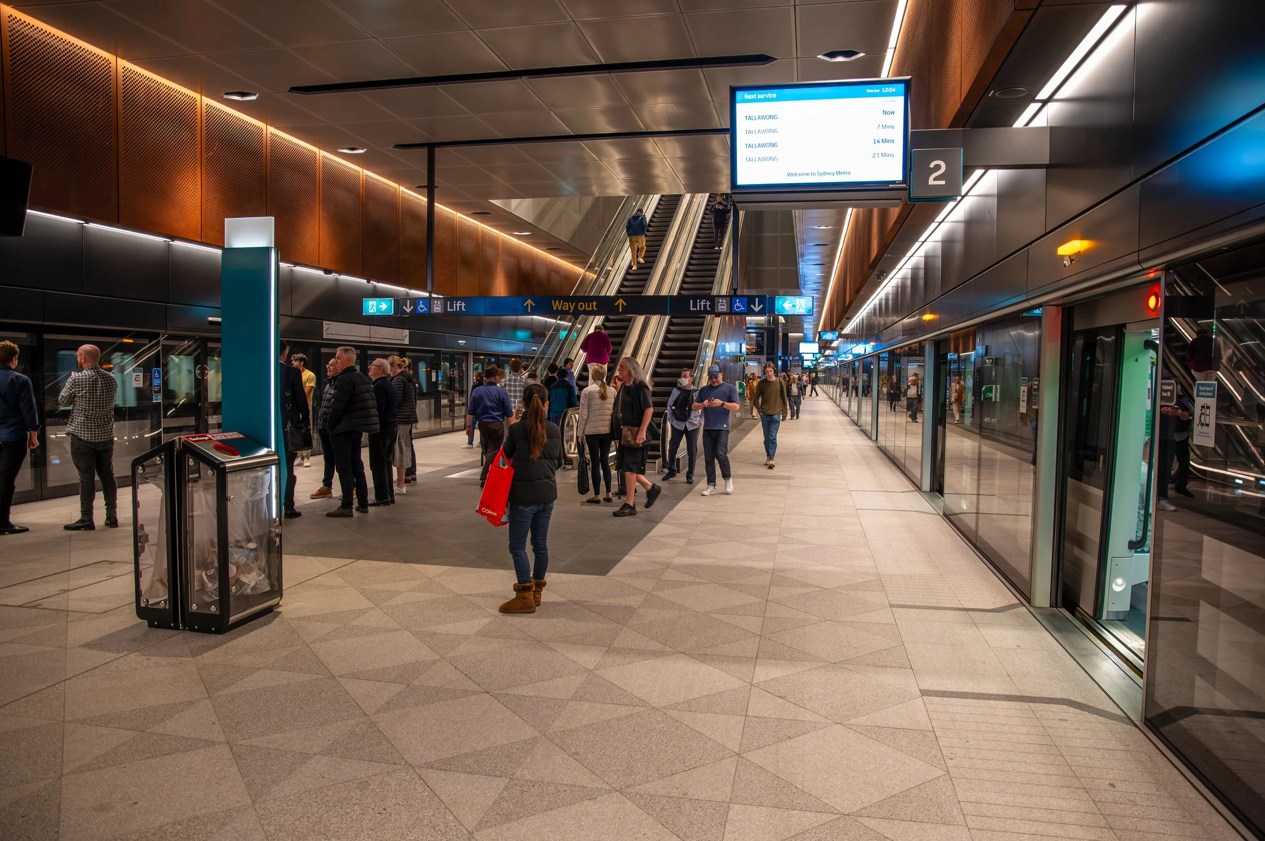 Interior of a modern subway or train station with people waiting and walking, escalators leading up, digital signboards displaying schedules, and glass doors.